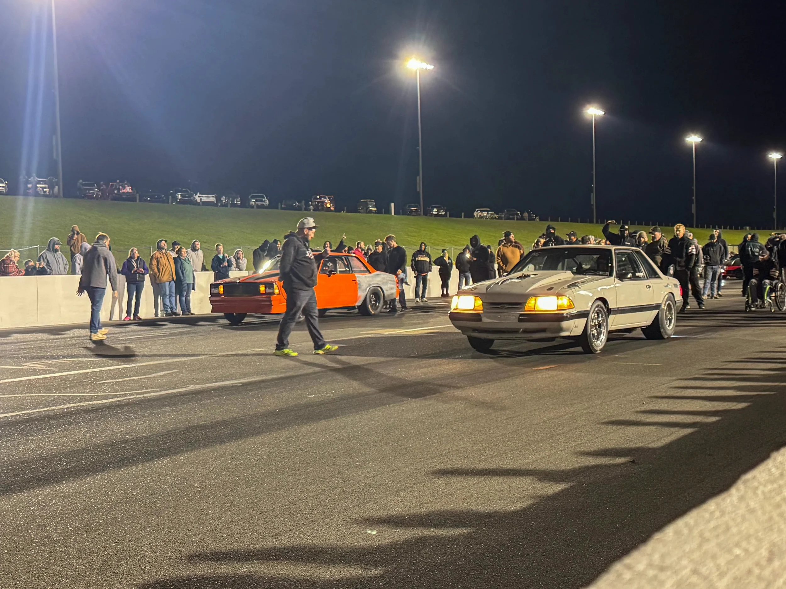 Nighttime scene in a parking lot with a crowd of people gathered along a barrier, classic cars including a red and black car, and multiple bright stadium lights illuminating the scene.