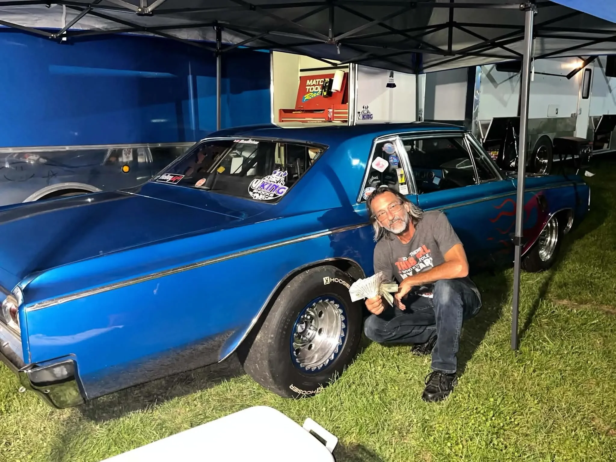 A man kneeling beside a blue vintage racing car with racing stickers on the window, under a canopy at a car show. There is a red tool chest and a white trailer in the background.