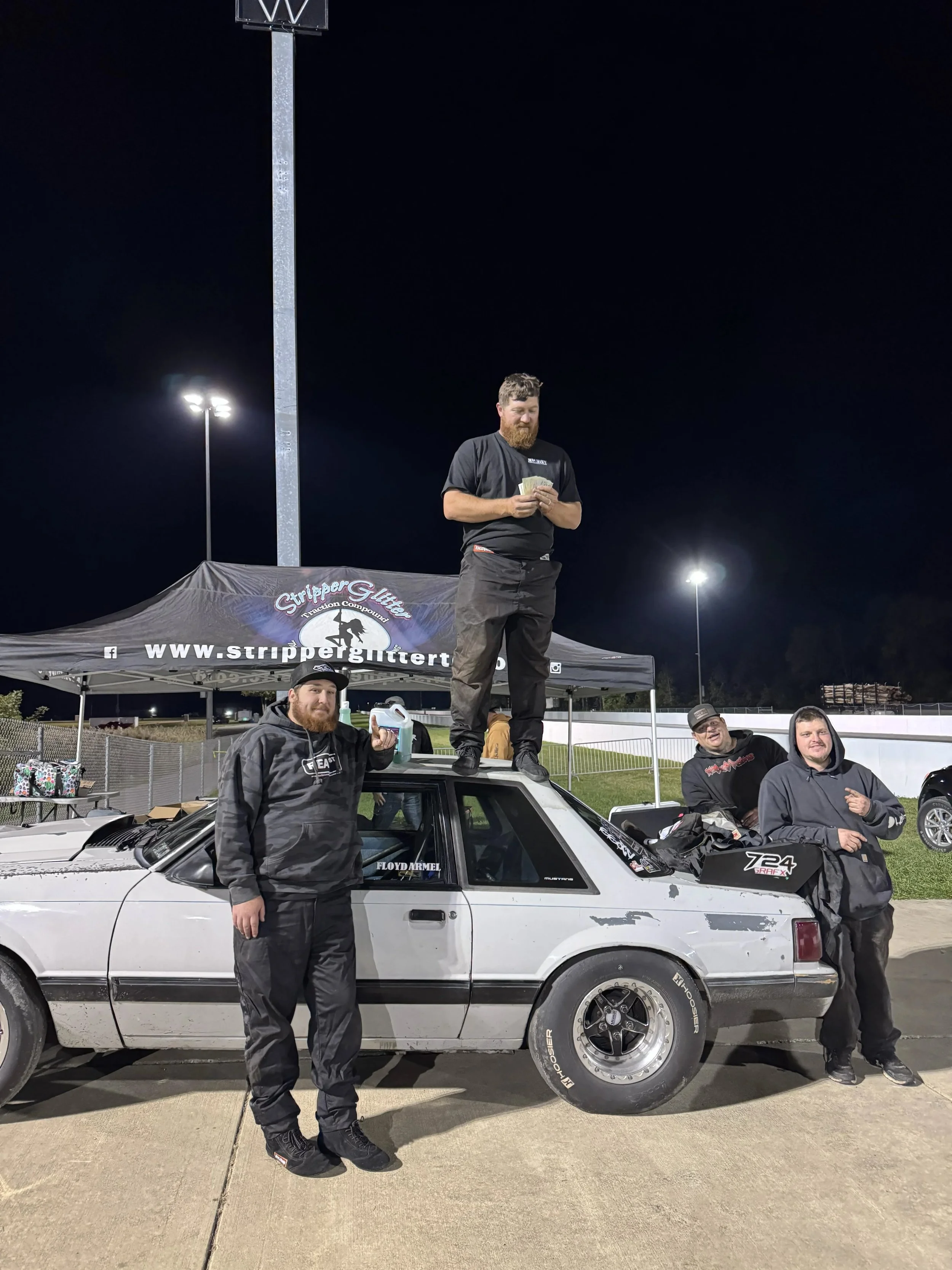 Four men at a night racing event standing around a white race car with a roof-mounted car load and a man standing on top of the car holding money, with a backdrop of a tent, a fence, and tall lights in a parking lot.
