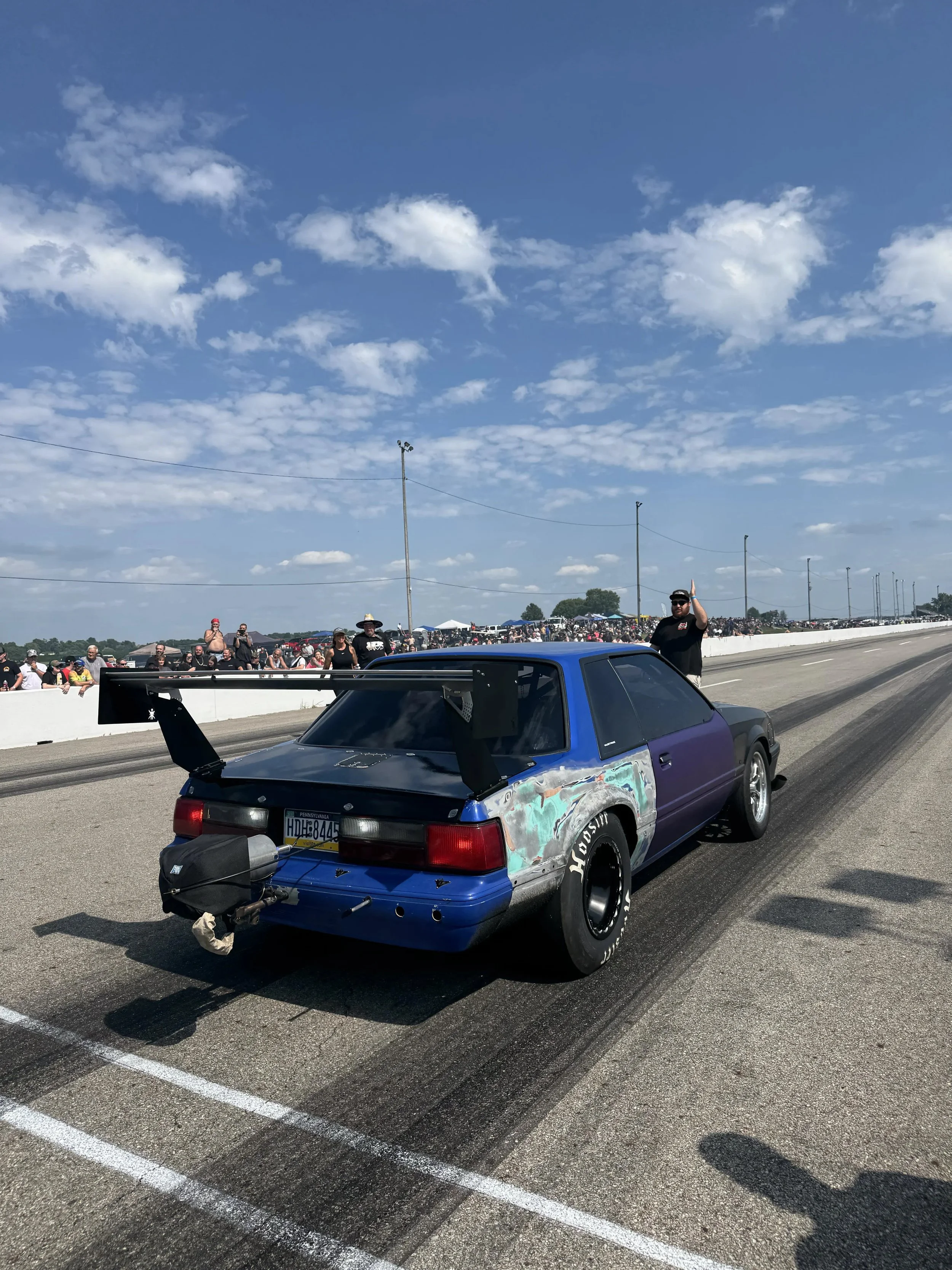 A blue race car on a racing track, with people and tents in the background and a partly cloudy sky overhead.