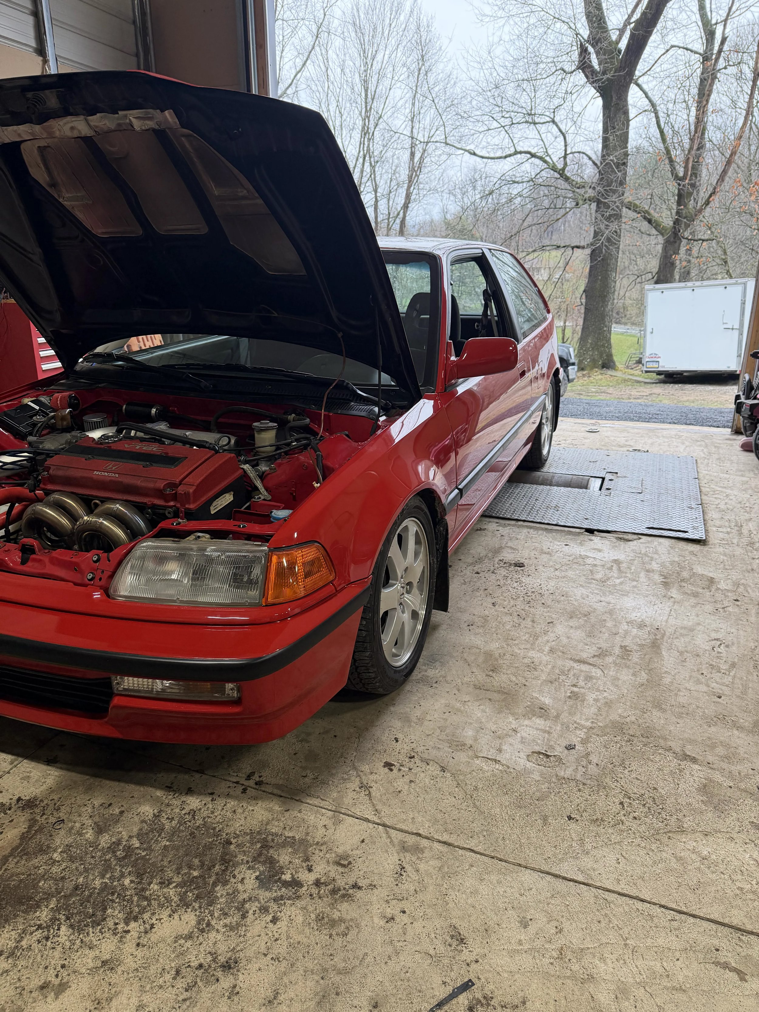 A red Honda Civic car with its hood open inside a garage, revealing the engine. The garage has a concrete floor and open garage door. Outside, there are trees and a lawn.