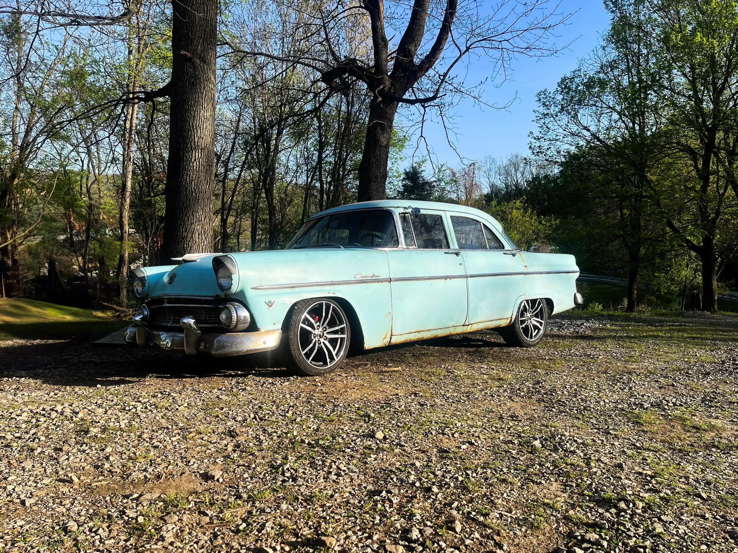 A vintage light blue car parked on a gravel area surrounded by trees with a clear blue sky above.