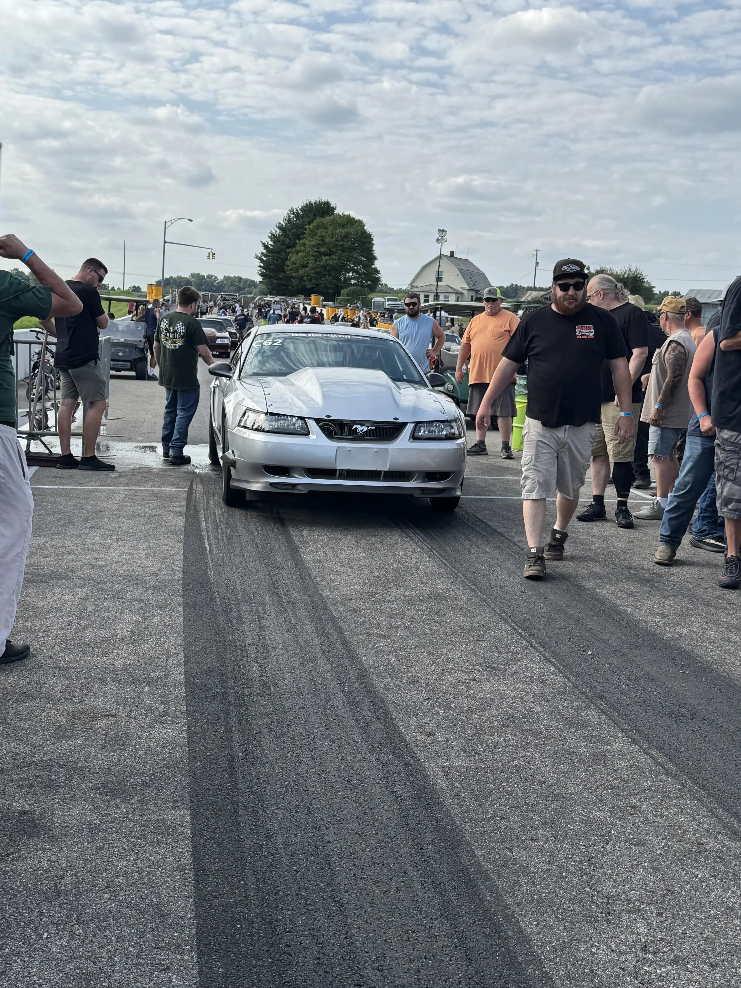 A silver car at a drag racing event with a group of people gathered around, some inspecting the car and others standing and talking. The car has a sticker on the windshield that reads 'Nitro Drag Week 2023' and the number 'C62'.