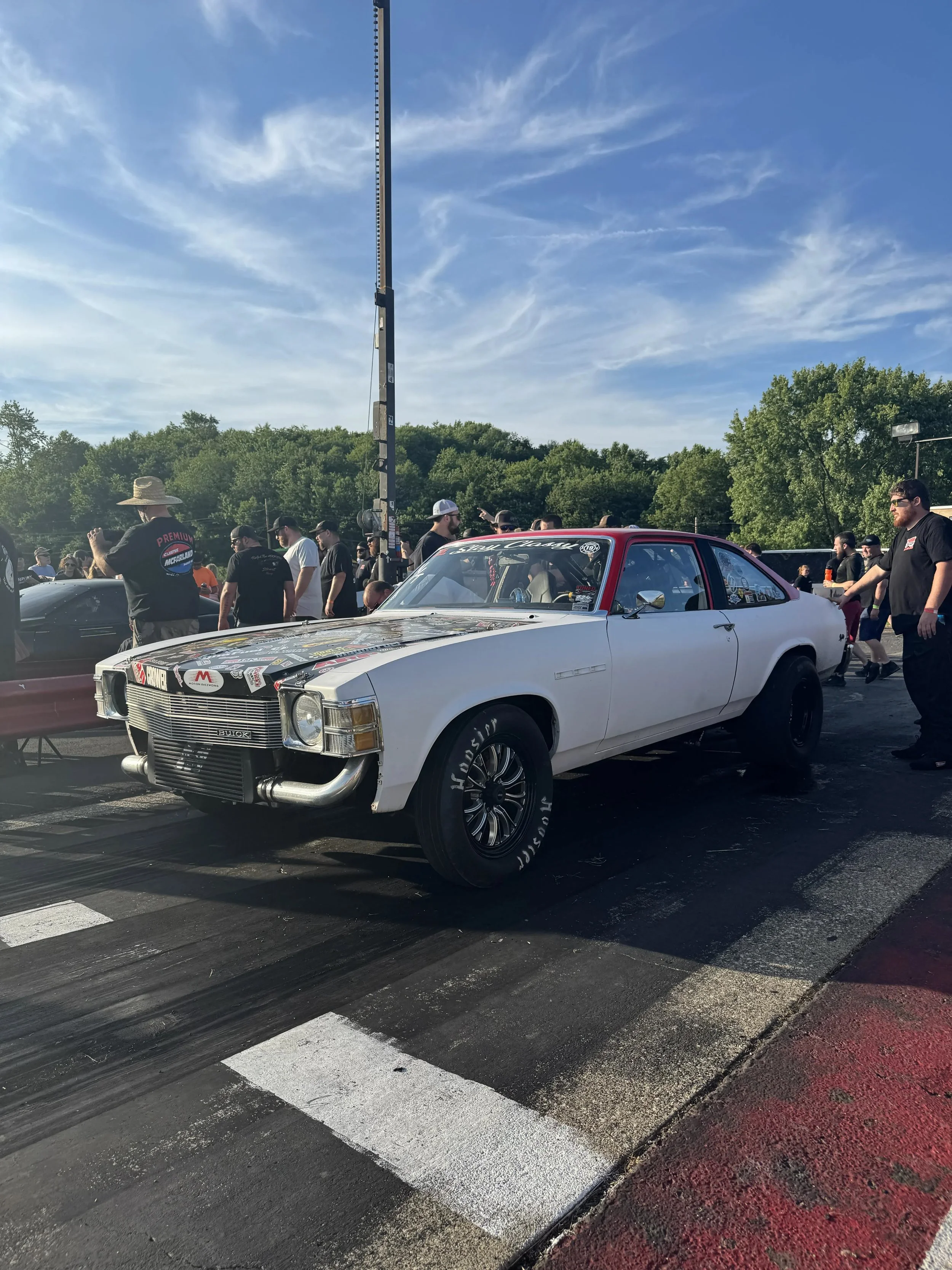A white vintage Buick car with a black hood and various stickers on it, parked at a car meet with people gathered around, against a backdrop of green trees and a blue sky with wispy clouds.