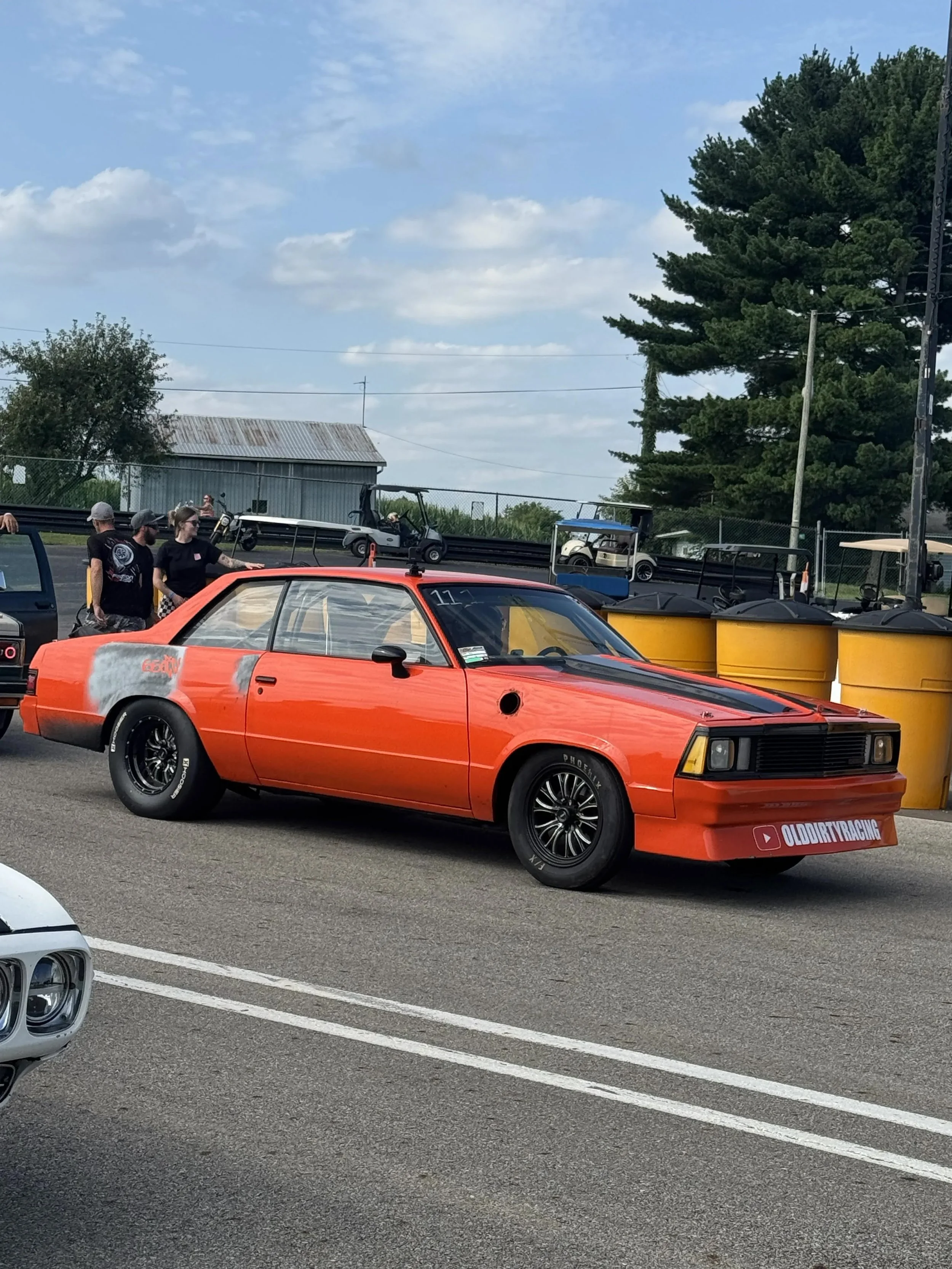 A red vintage race car with black wheels, parked in a lot, with a sticker that reads 'OLDIRTYRACING' on the front bumper. There are people standing nearby and some golf carts and other vehicles in the background, with trees and a building under a par