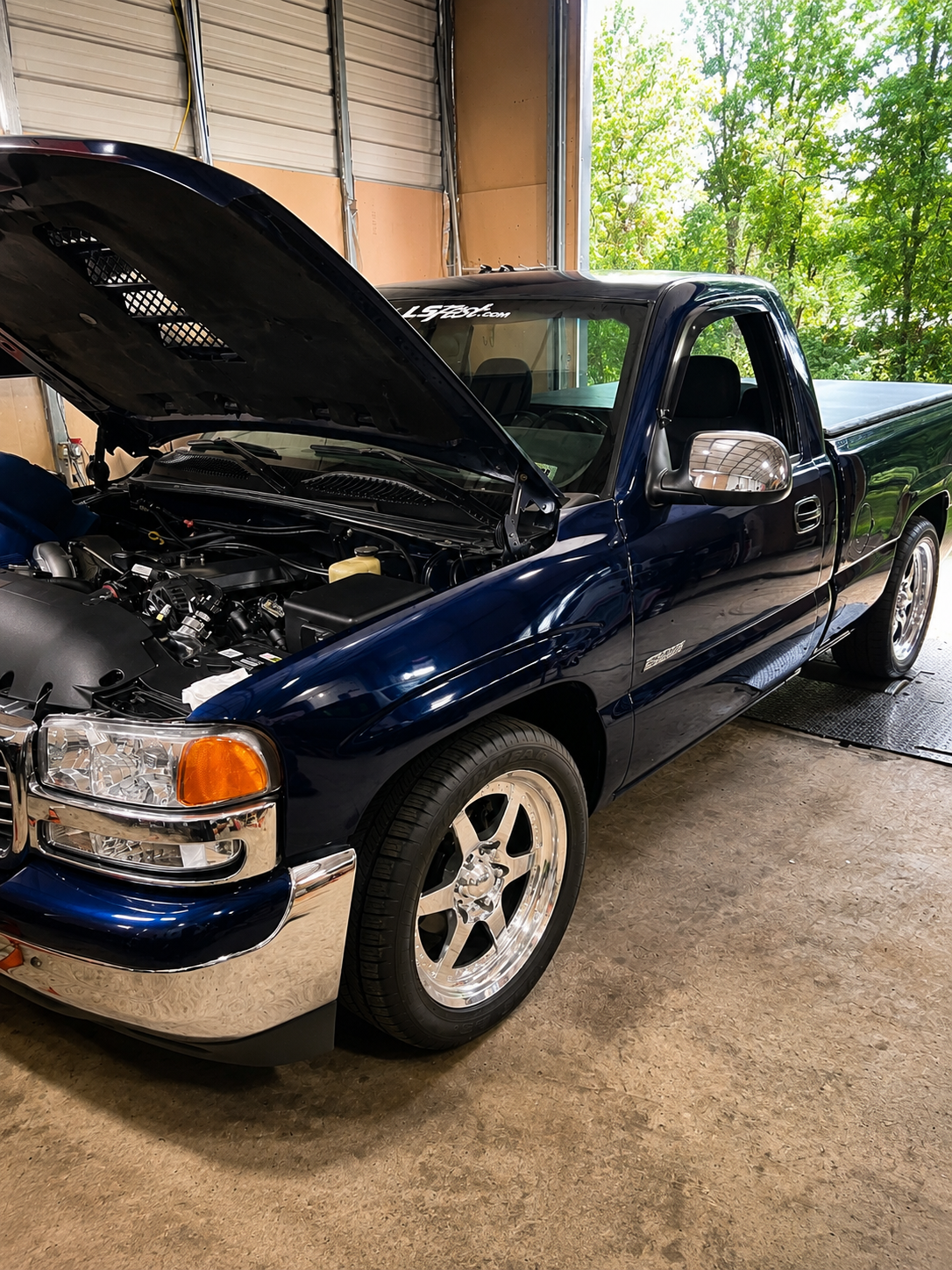 A dark blue pickup truck with the hood open inside a garage, revealing the engine. The garage has an open door showing green trees outside.