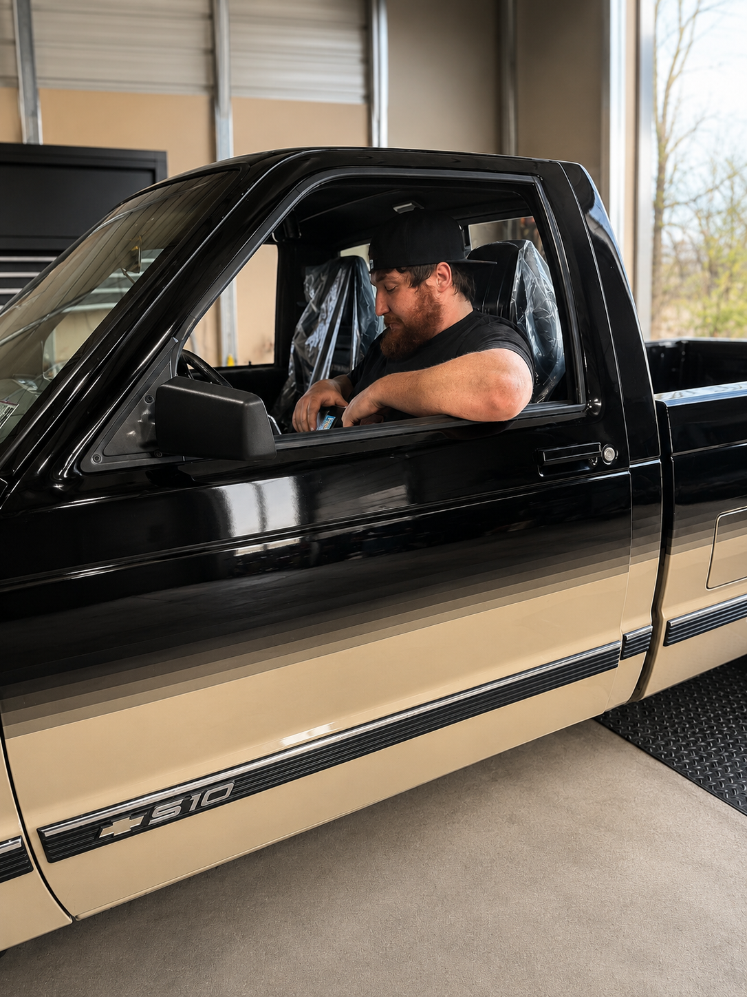 Man sitting in the driver's seat of a black and gold truck, looking at his phone inside a garage.