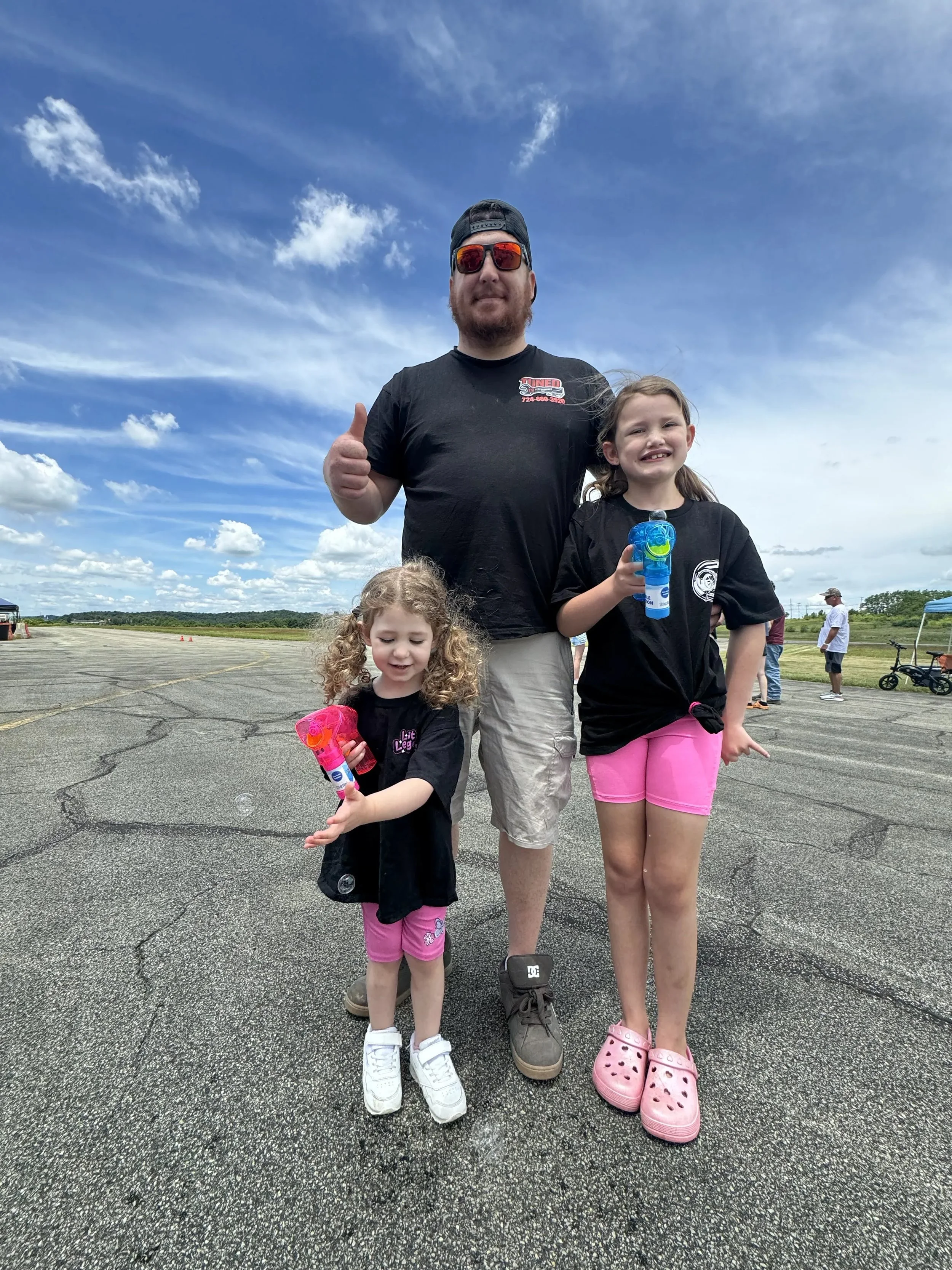 A man with a beard, sunglasses, and a black cap giving a thumbs-up, standing with two young girls holding bubble wands outdoors under a partly cloudy sky.