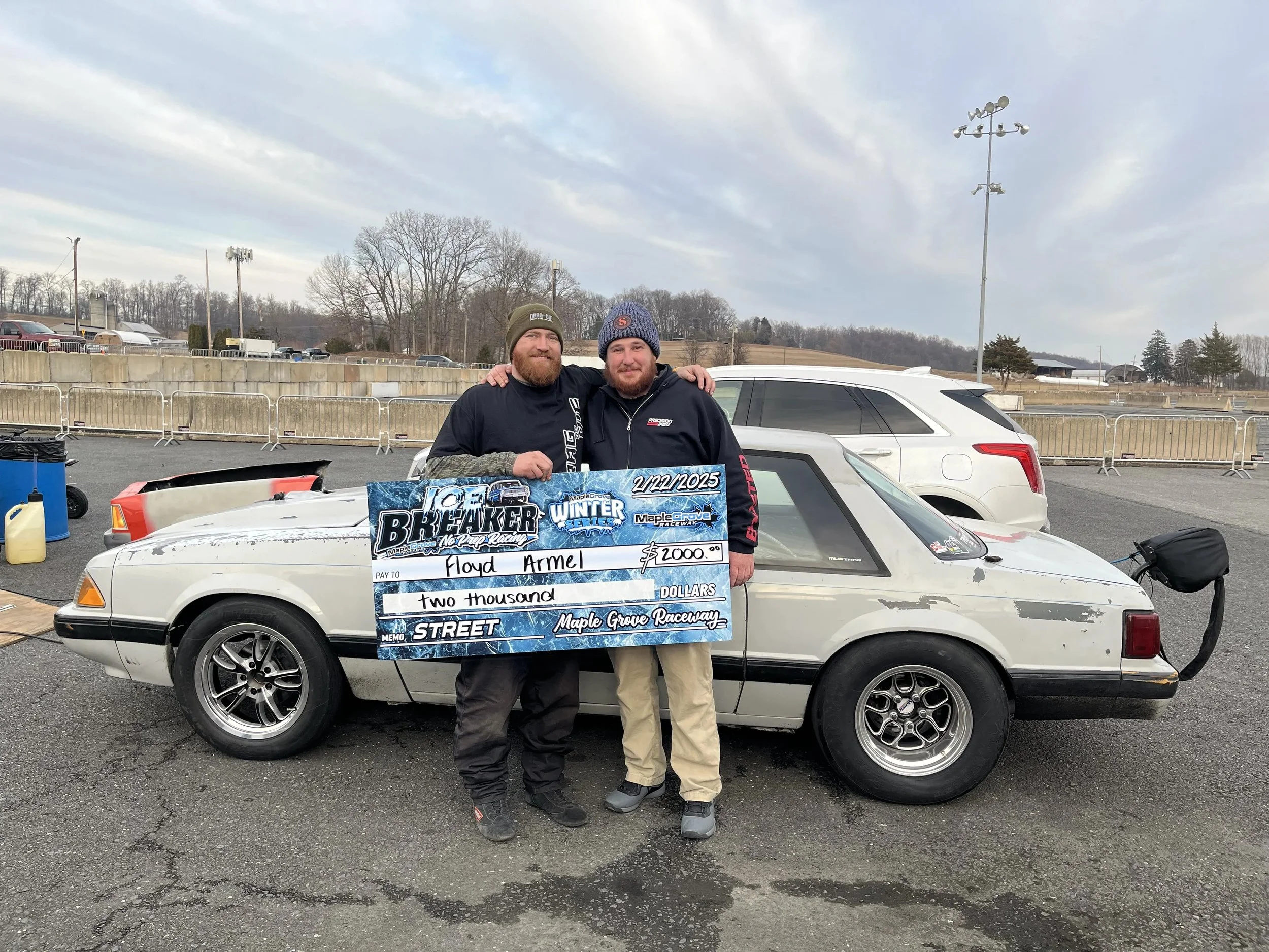 Two men standing together outdoors, holding a large check for $2,000. The check is made out to Floyd Armell and is dated February 22, 2025. They are smiling and wearing winter clothing, with one man wearing a brown beanie and the other a blue beanie,