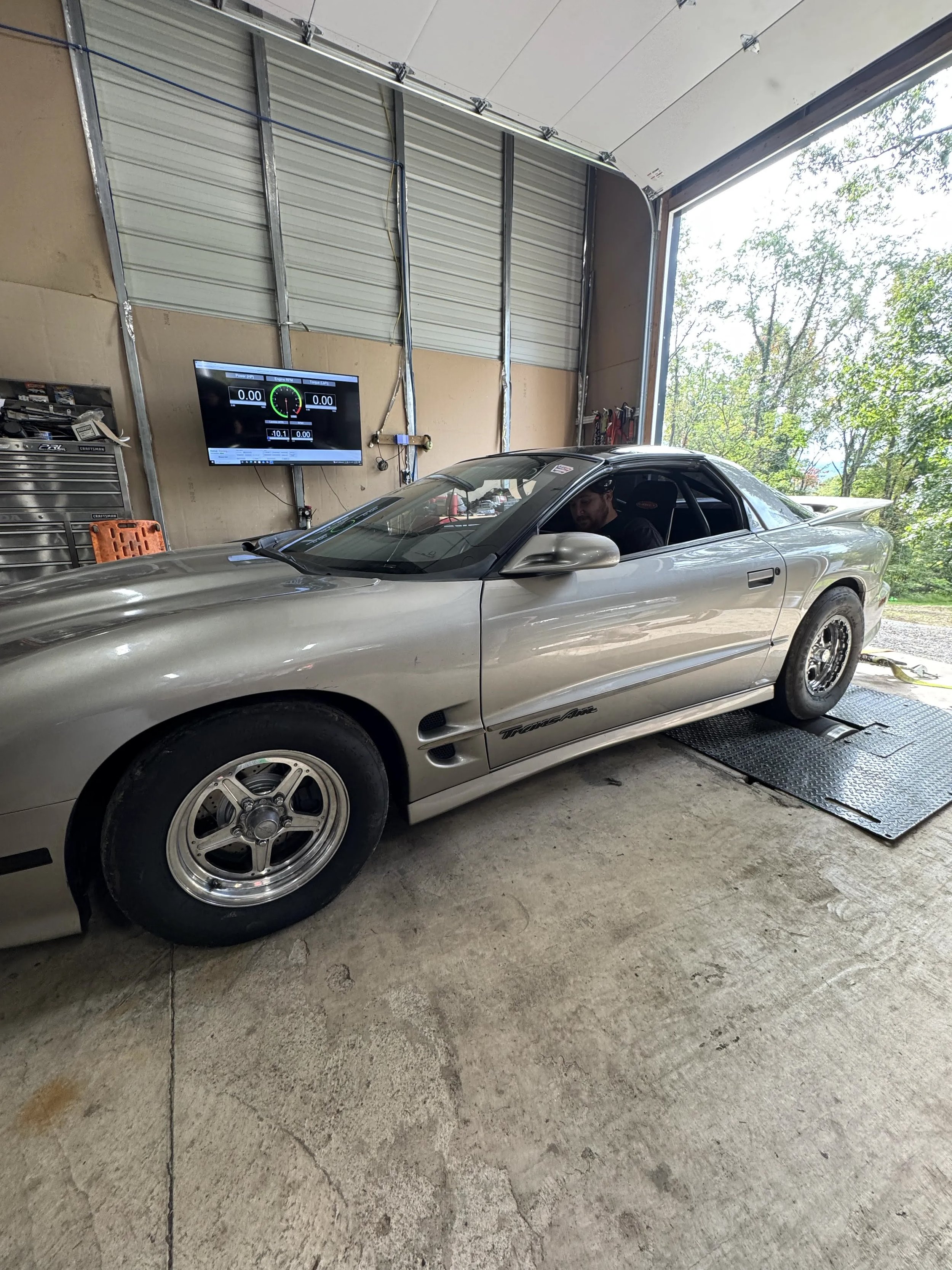 A silver sports car inside a garage, with a person sitting in the driver's seat. The garage has an open door showing trees outside, a large wall-mounted screen, and some tools on the wall.