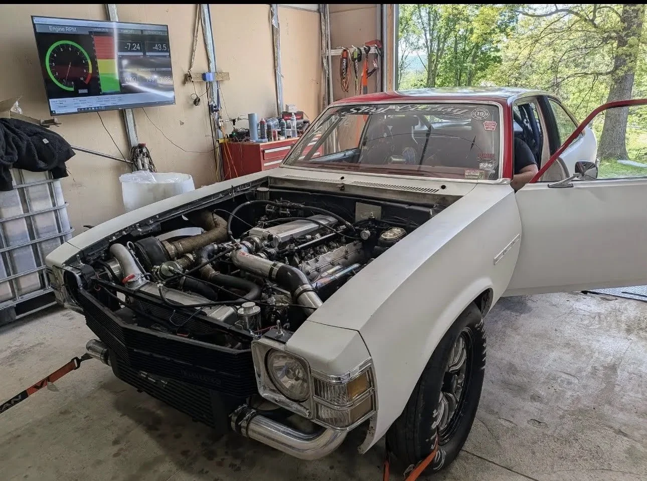 A classic muscle car in a garage with its hood removed, exposing the engine. The car has a white exterior, a red interior, and slick black wheels. The garage has tools, bottles, and a large digital gauge display on the wall, with trees visible outsid