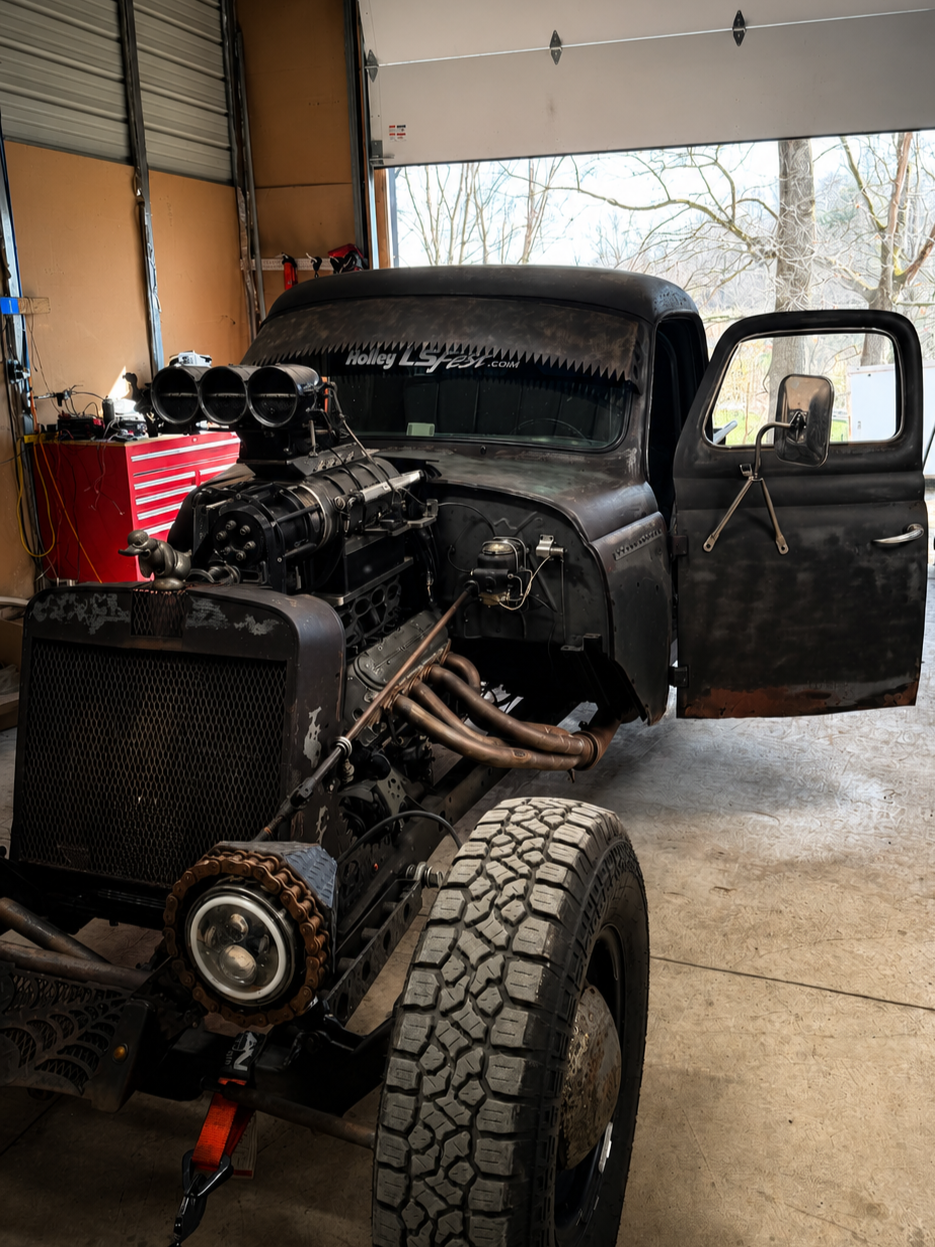 A vintage black truck with a large engine exposed, inside a garage. The truck has no front bodywork, large off-road tires, and a windshield with a sticker that says 'Holley LS Fest.com.' The garage has a red tool chest and some tools on the wall. A garage door is open, revealing trees outside.