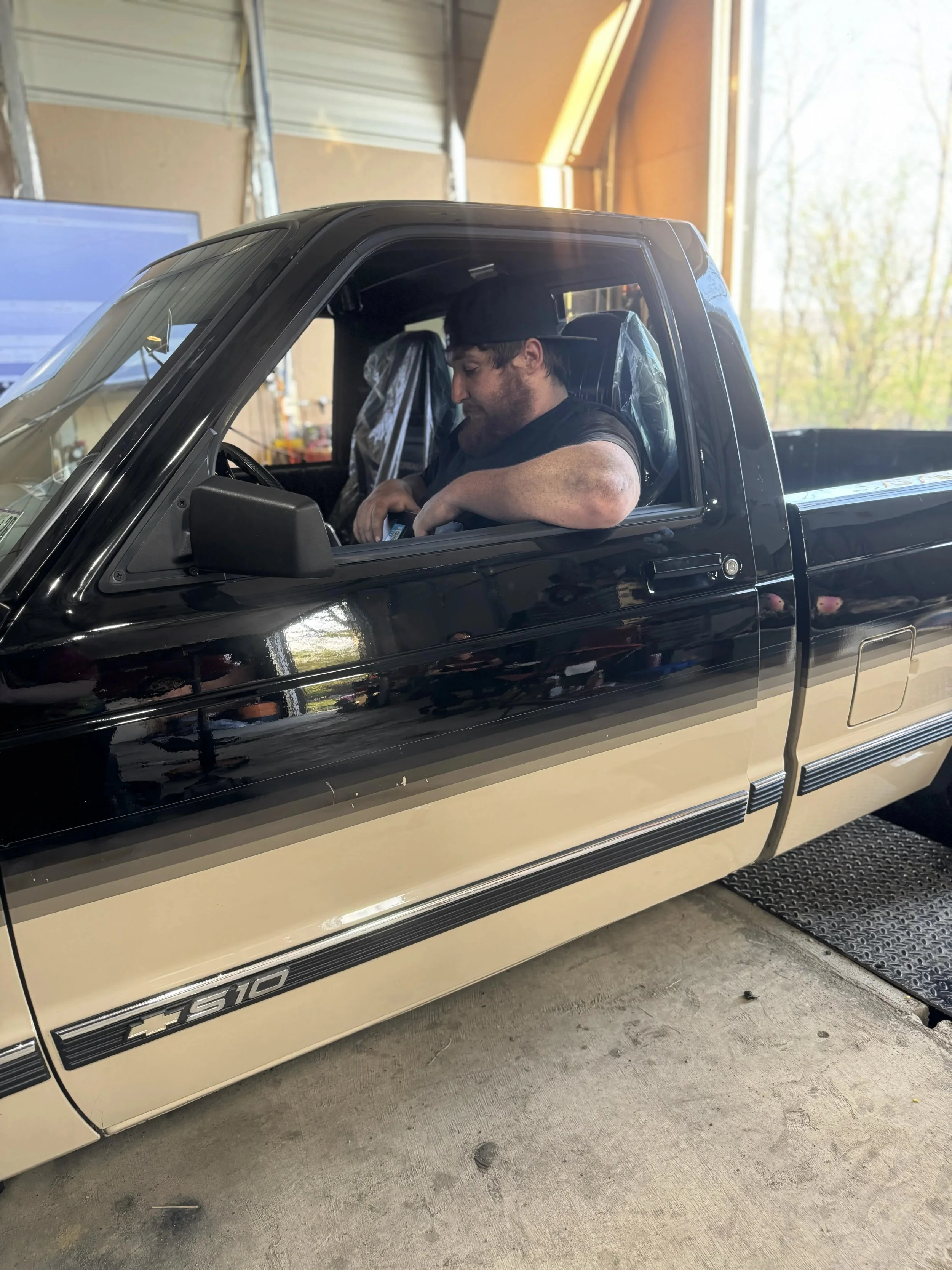 A young man with a beard and a black cap sitting in the driver's seat of a black and beige pickup truck inside a garage. The garage has a computer monitor and tools in the background and is illuminated by natural light from an open door.