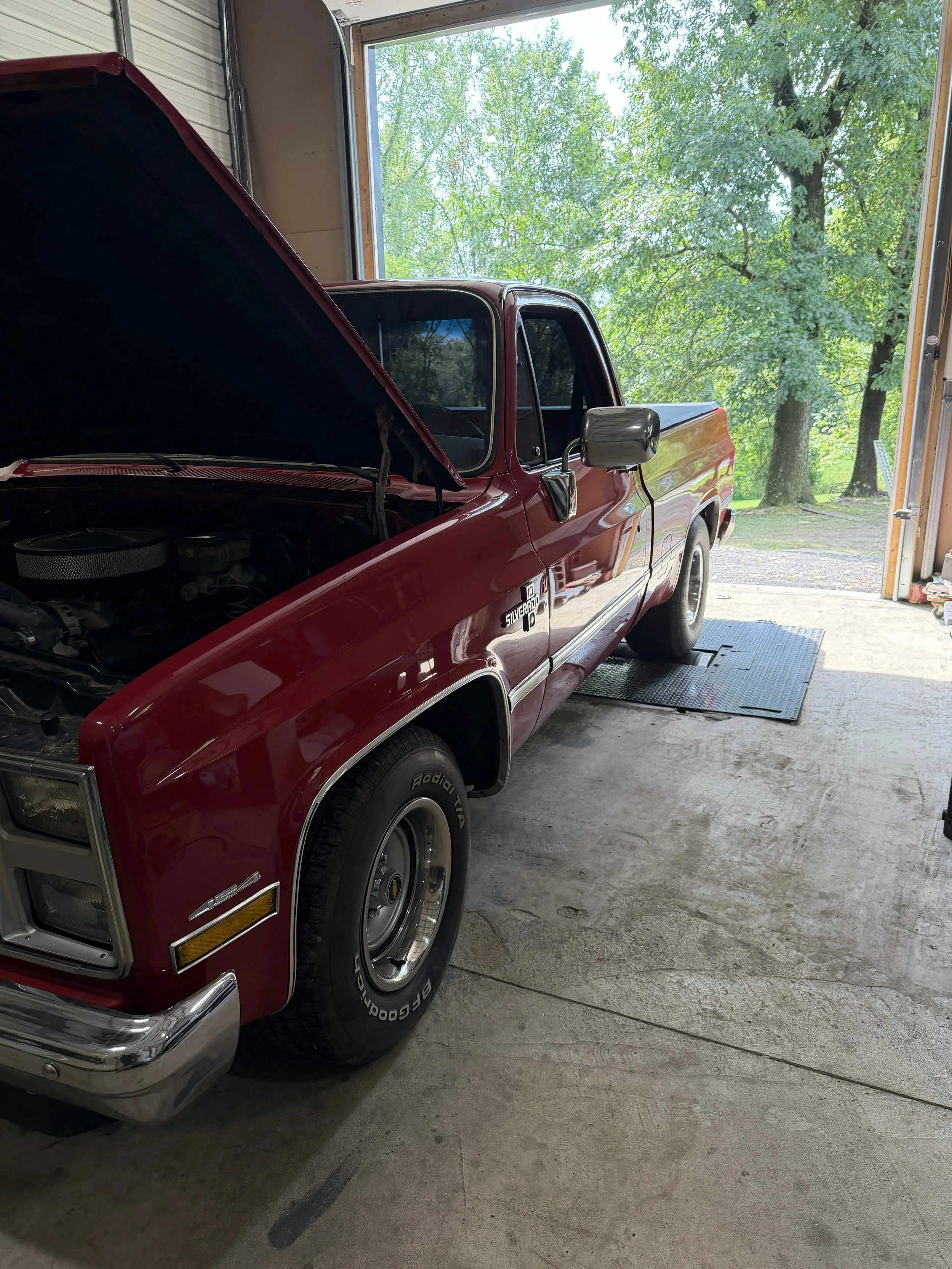 A red Chevrolet Silverado pickup truck in a garage with its hood open, showing part of the engine, with a large open garage door revealing green trees outside.