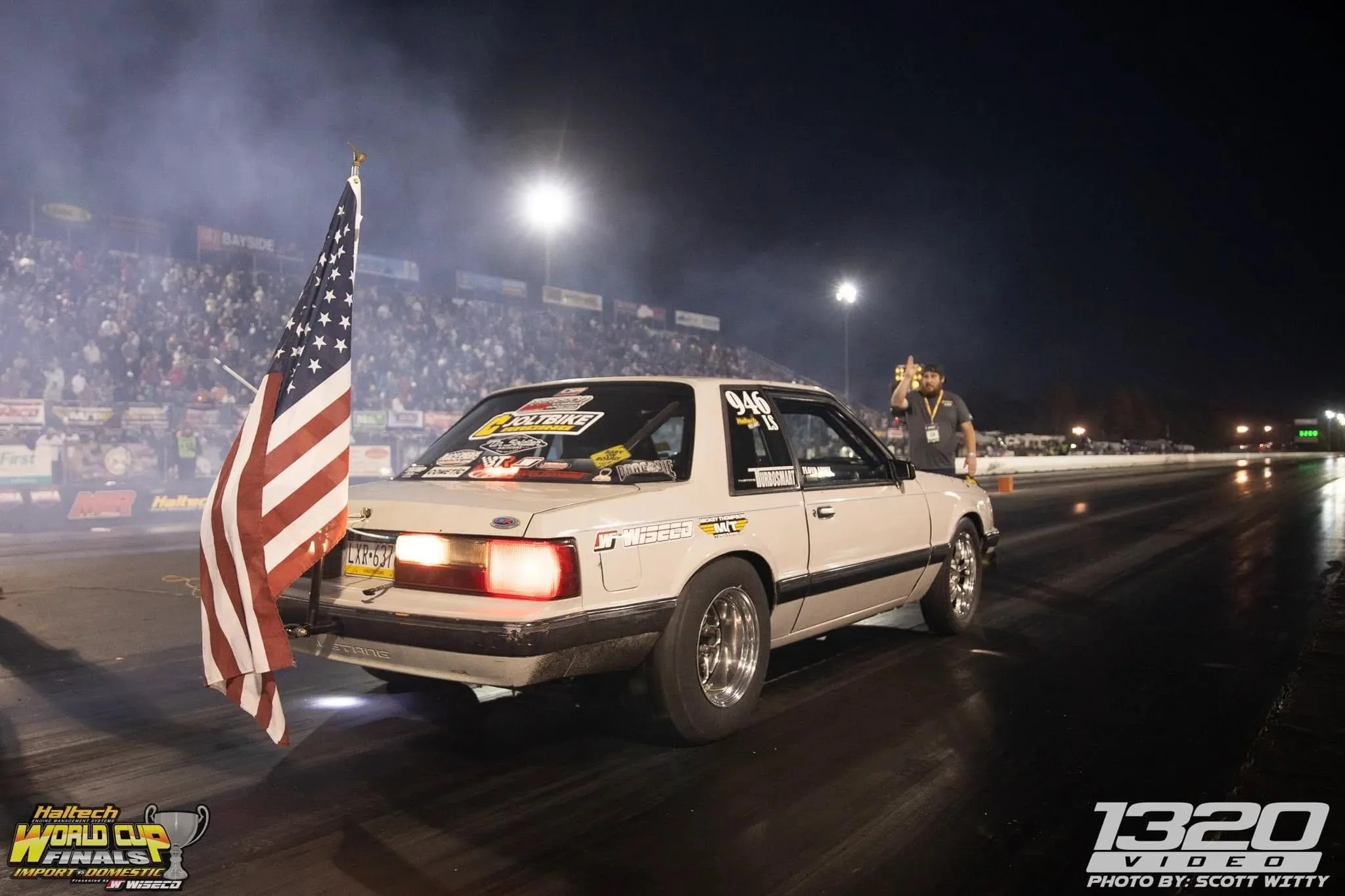 A white racing car with stickers on the rear window, positioned on a drag strip at night, with an American flag attached to a pole in the foreground and a crowd in the background. Bright lights illuminate the scene, and a person stands beside the car