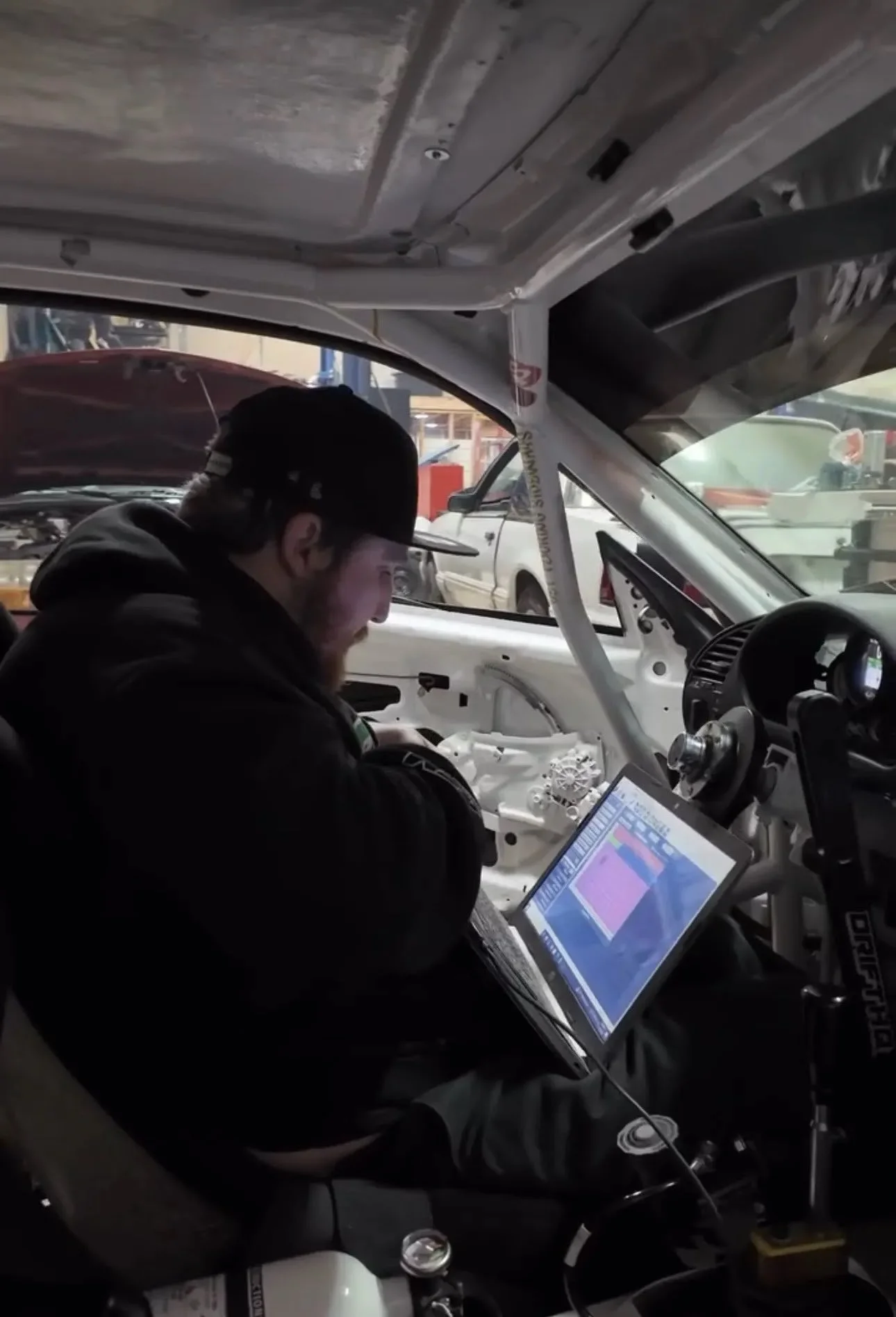 A person working inside a race car with a partially assembled interior, using a laptop and other electronic equipment, in a garage or workshop.