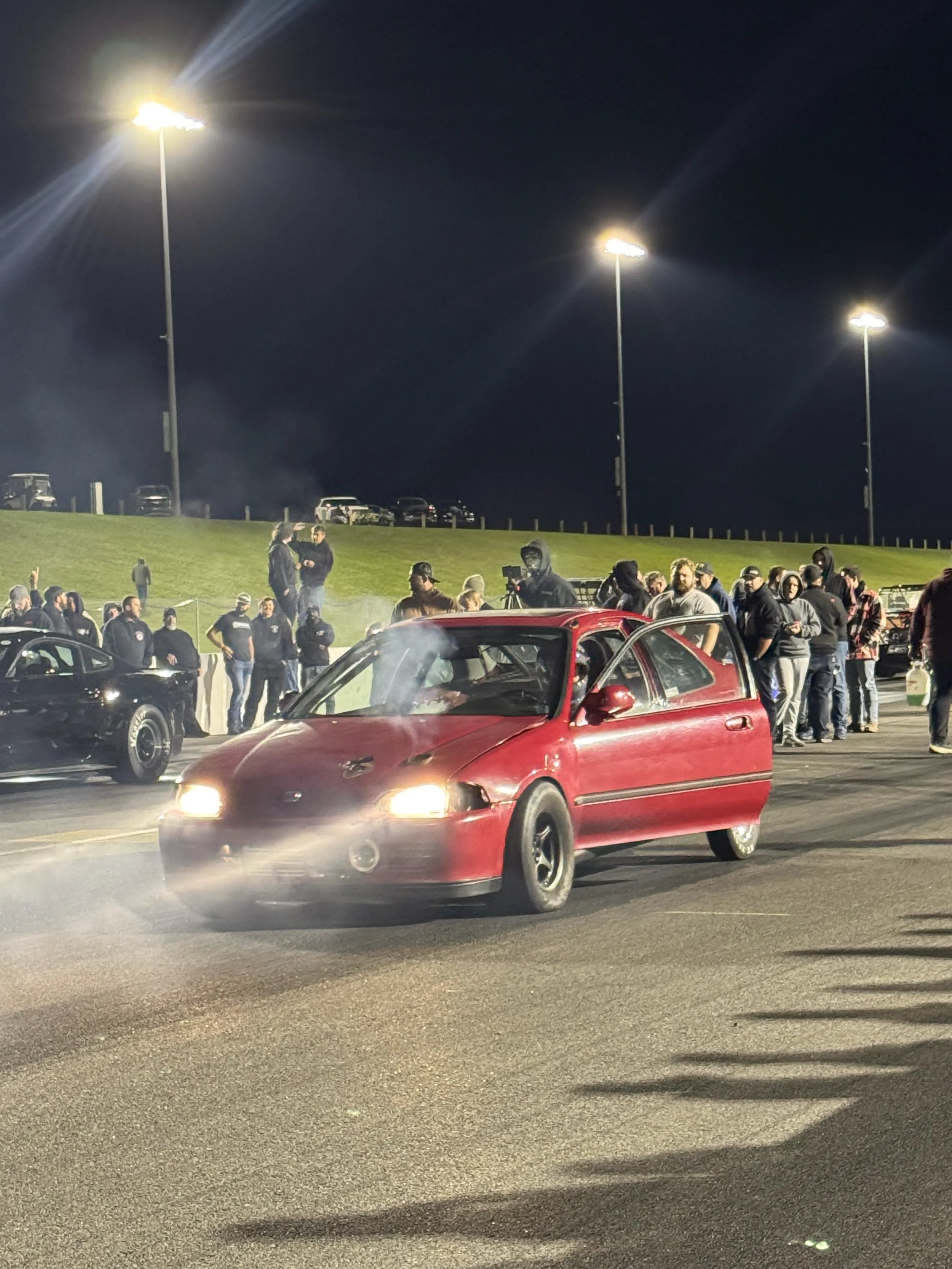 Nighttime scene of a car meet or racing event with several cars parked and people gathered. Bright streetlights illuminate the area, and a red sports car is in motion on the road in the foreground.