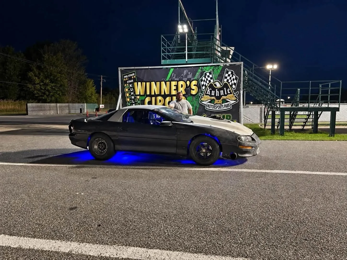 A black race car with blue underglow lights is parked in front of a sign for Kuhnla Motorsports Park at night. A person is standing behind the car. The background includes trees and a race track with orange cones.