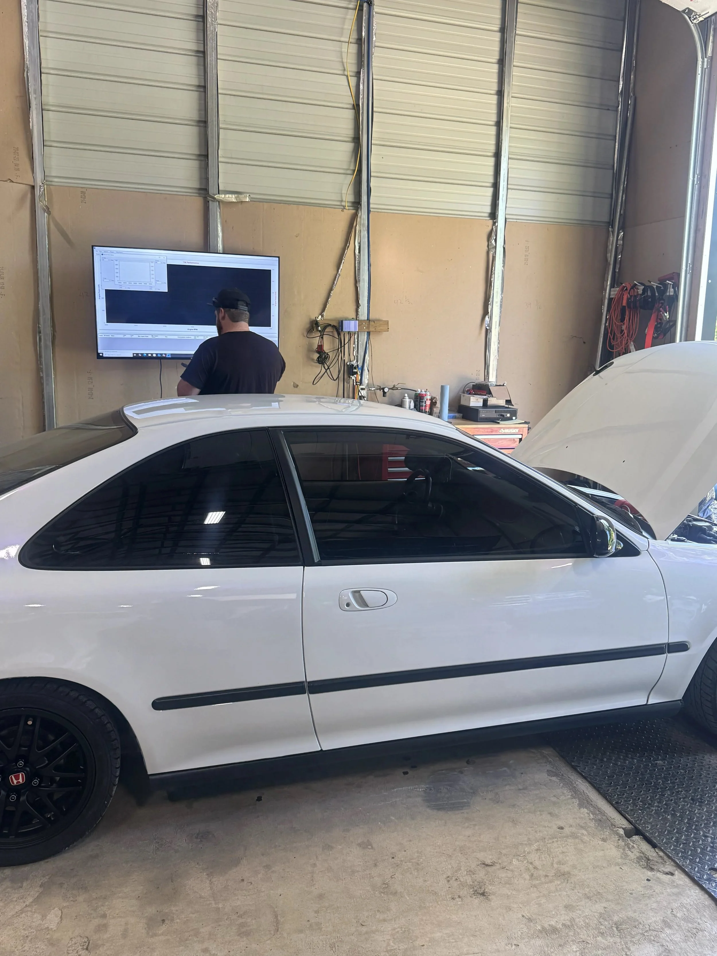 A white car with its hood open parked inside a garage, with a person standing next to a large screen on the wall.