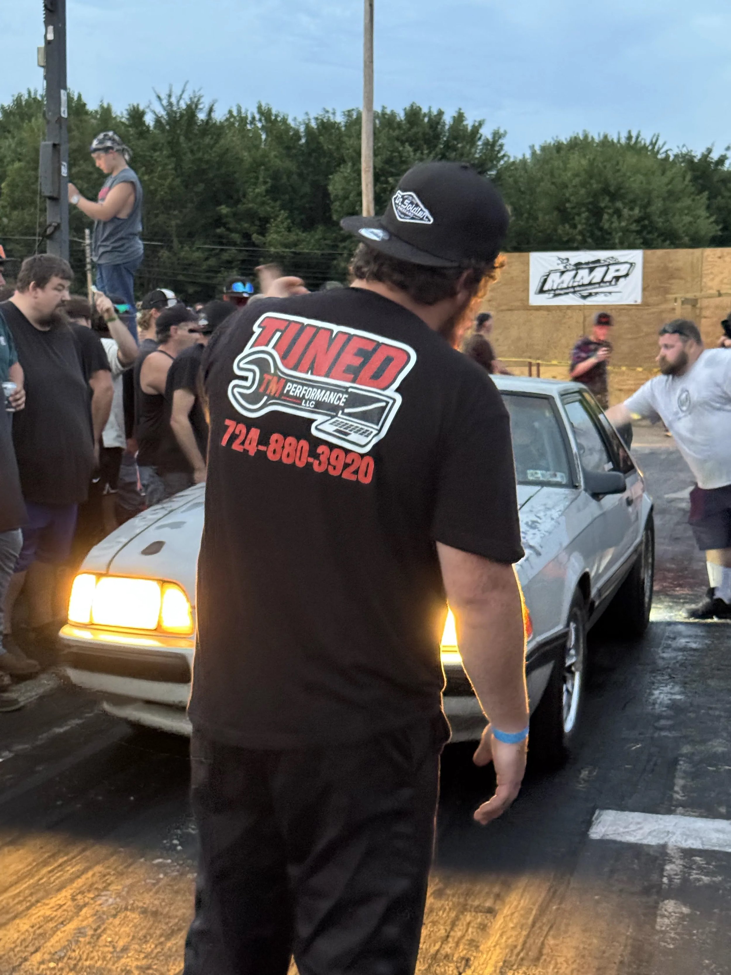 A crowd at a racing or car event, with people standing around a vintage silver car, some using phones. A man in the foreground wears a black t-shirt with a logo and the word 'TUNED' and the phone number 724-880-3920. In the background, there are tree