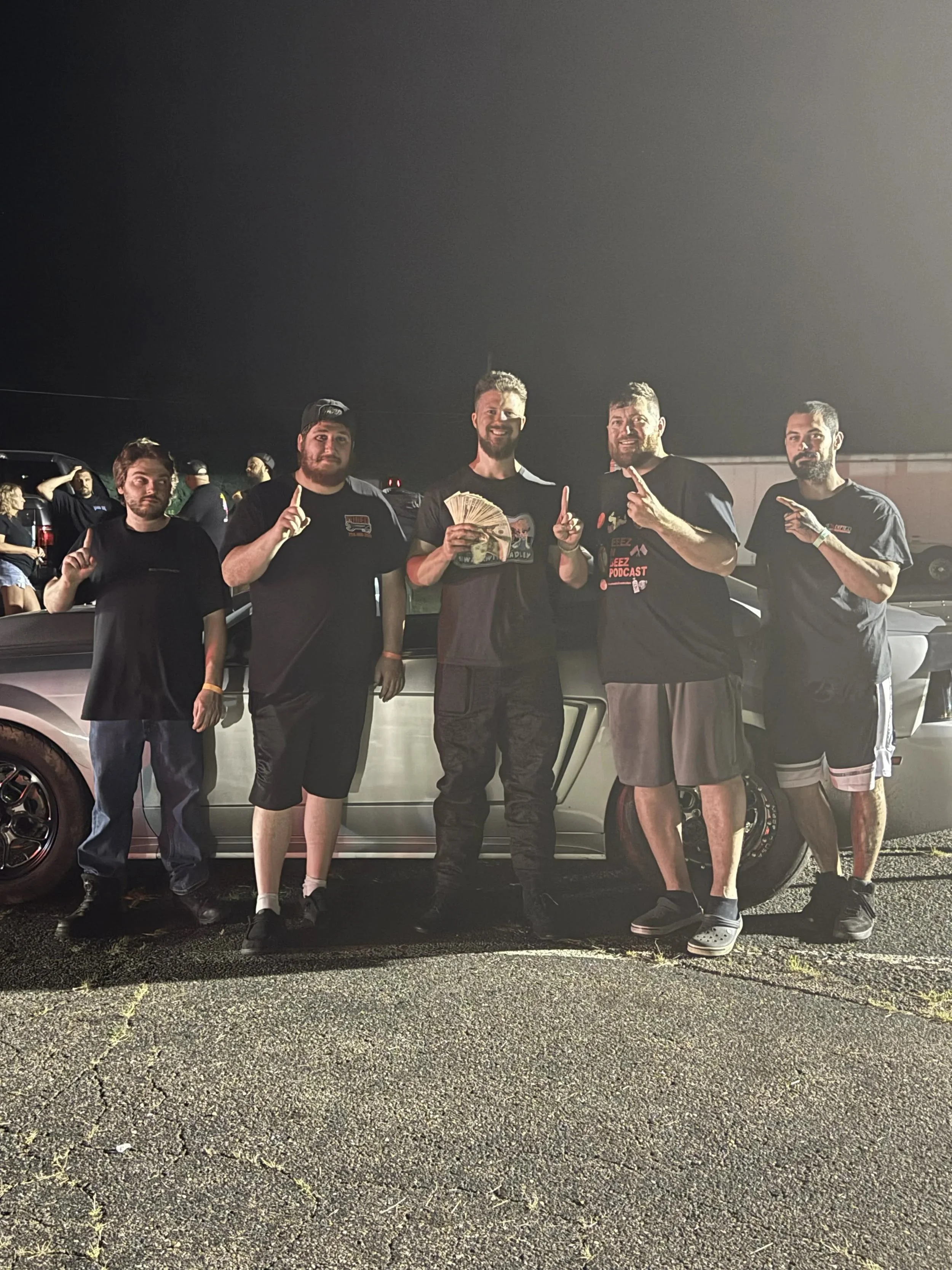 A group of five men standing outdoors at night, some holding their index fingers up, with a silver car behind them and other people visible in the background.