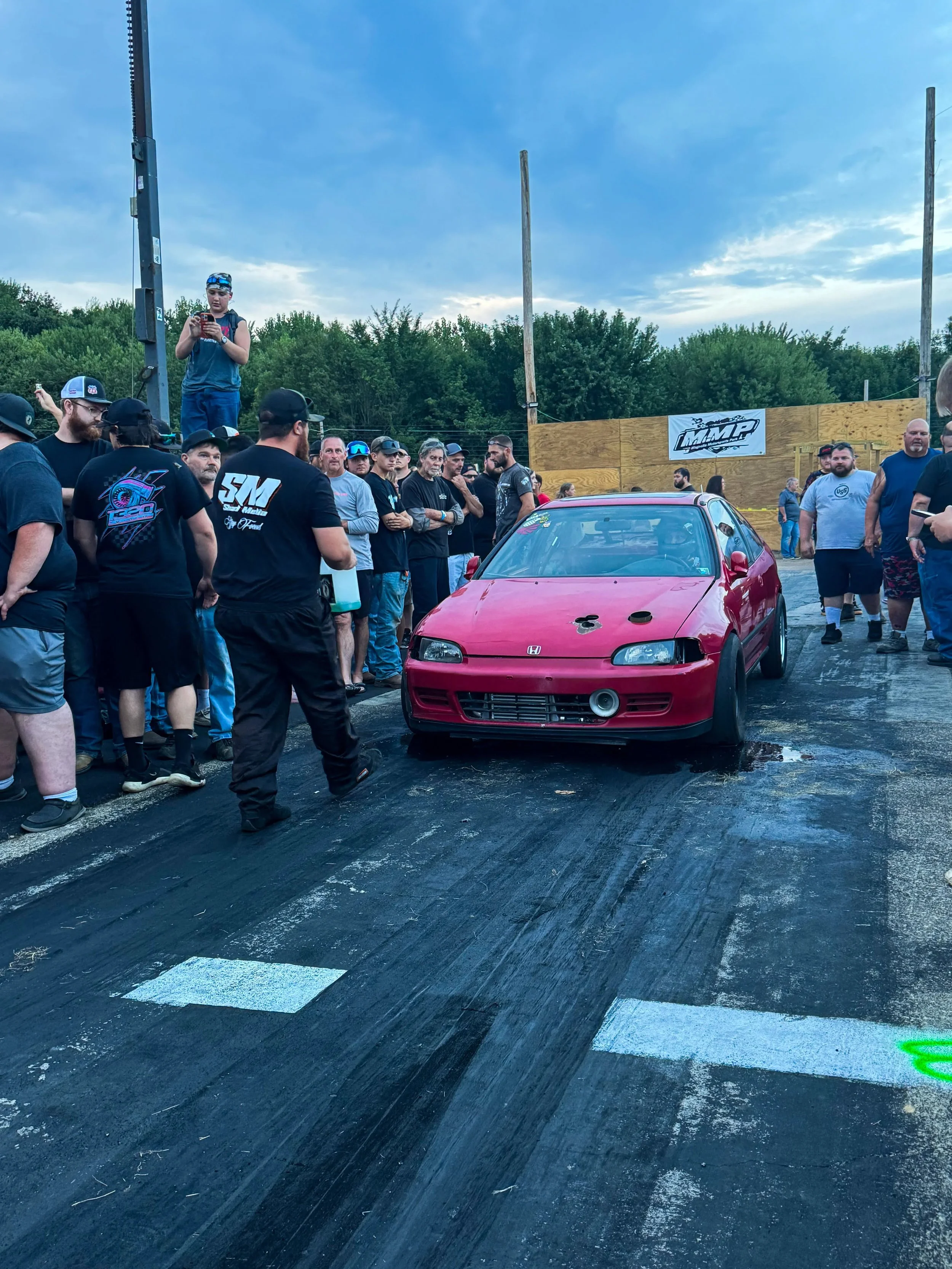 A red car is at a drag racing event surrounded by a crowd of spectators and participants. The car has a damaged front bumper and hood. There are people watching and taking photos, and a person standing on a platform with a phone. The scene takes plac