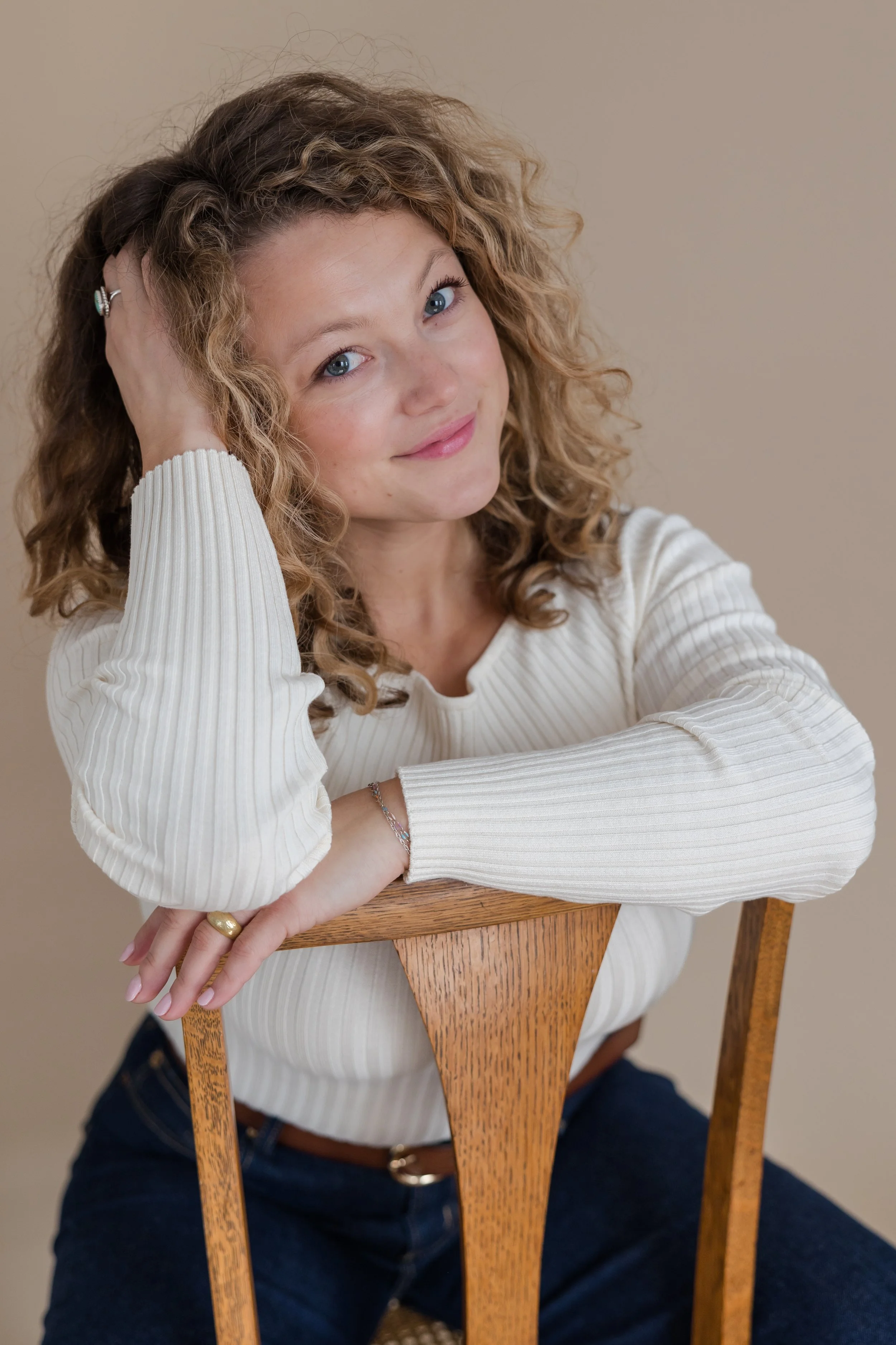A young woman with curly blonde hair, blue eyes, and fair skin sitting on a wooden chair, leaning on the backrest with her left arm resting on it, smiling and looking at the camera. She is wearing a white, long-sleeve, ribbed shirt and dark blue jeans.