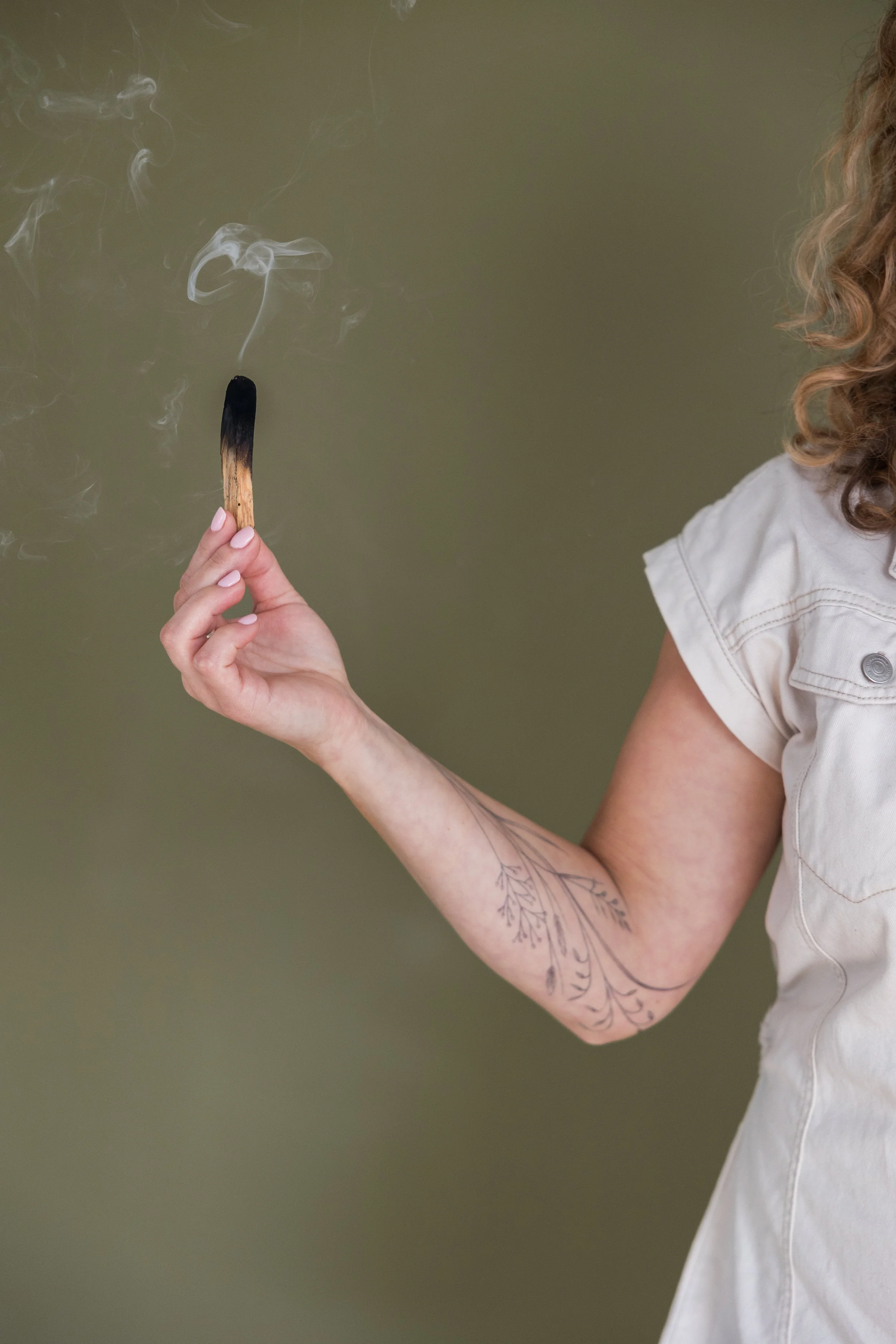 Person holding a lit matchstick with smoke rising from it, visible arm with a floral tattoo, wearing a short-sleeved white shirt, against a plain, muted olive-green background.