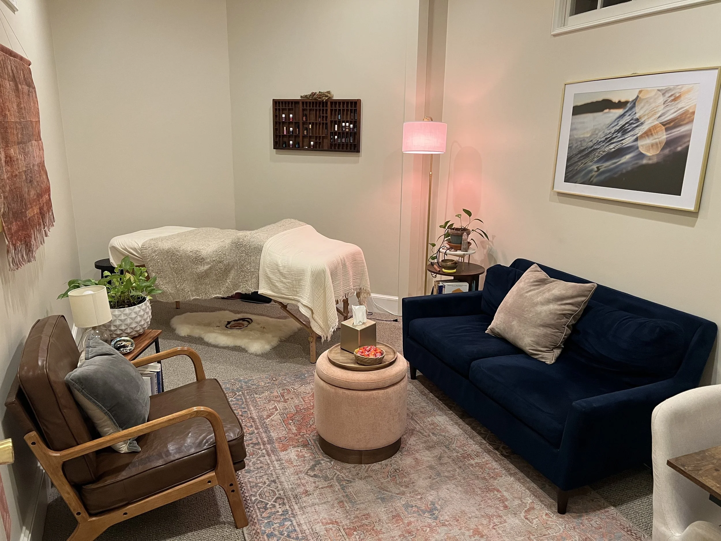 The image shows a cozy living room with a massage table covered in a white sheet and beige blanket, a navy blue sofa with beige pillows, a wooden chair with a gray pillow, a round ottoman with a bowl of potpourri, a small side table with a potted plant, a pink floor lamp, a framed mirror on the wall, a piece of wall art, a wall-mounted wooden spice rack, and a patterned area rug.