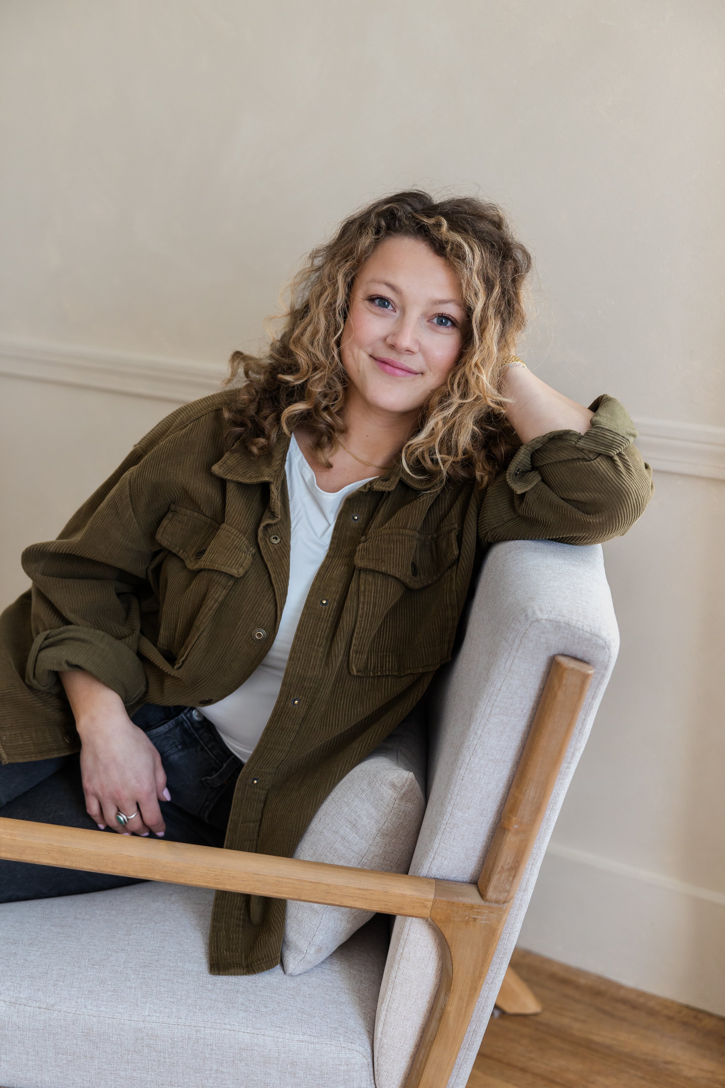 A woman with curly blonde hair sitting on a beige chair, resting her arm on the back of the chair and smiling at the camera.