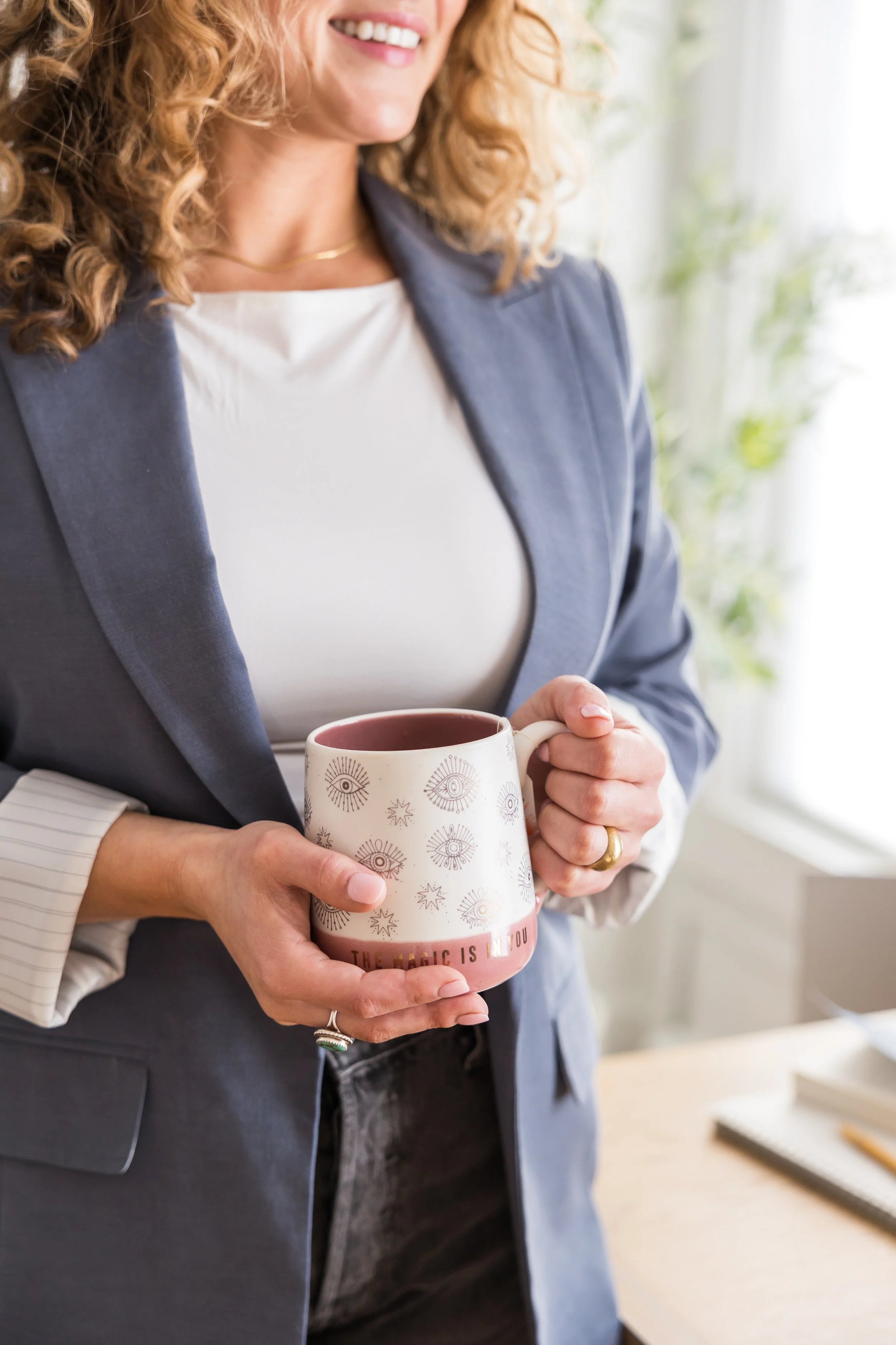 A woman smiling while holding a pink mug decorated with gold celestial designs, with another person’s hand also touching the mug. The woman wears a navy blazer over a white top and has curly red hair.