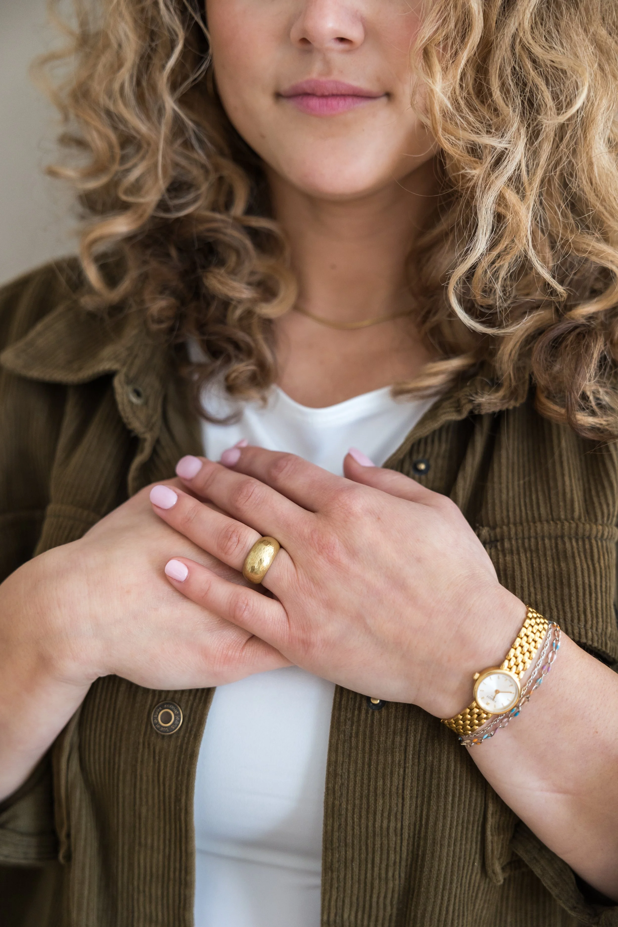 Close-up of a woman with curly blonde hair, wearing a white top and a green corduroy jacket. She has a gold ring on her finger, a gold watch, and multiple bracelets on her wrist. Her hands are crossed in front of her chest.