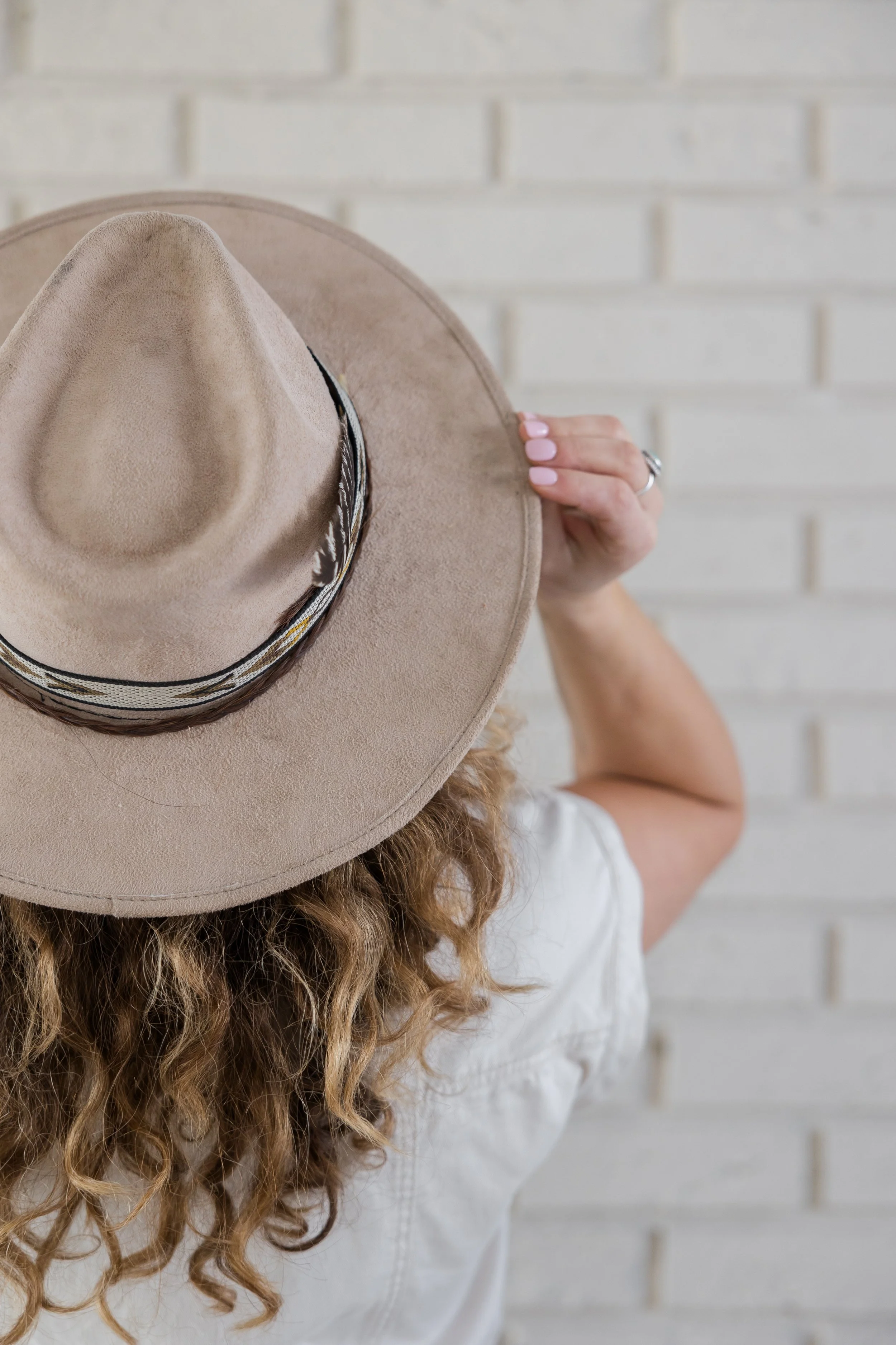 Person with curly hair holding a tan wide-brimmed hat with a patterned band, standing in front of a white brick wall.