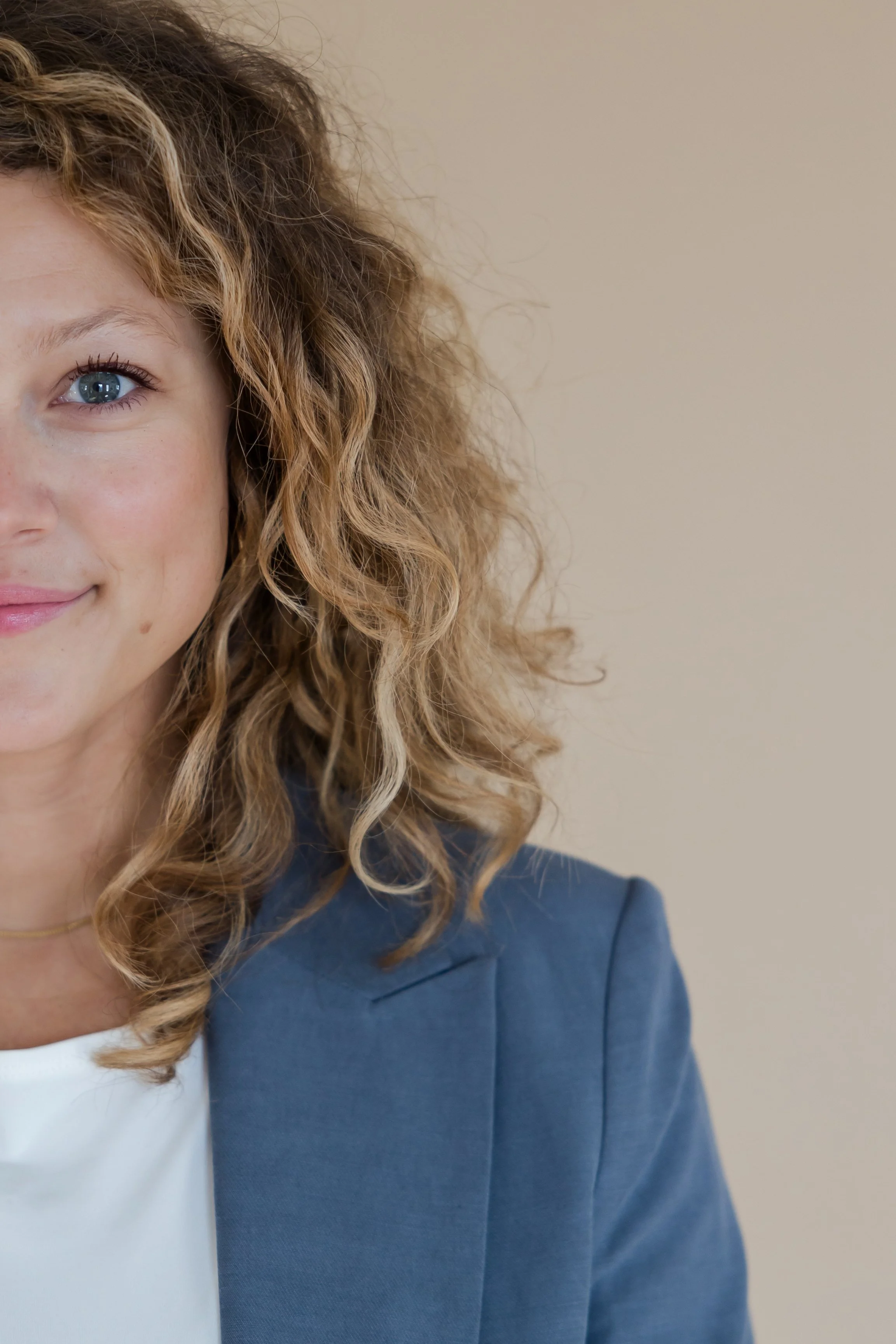 A woman with curly, blonde hair and blue eyes smiling, wearing a navy blazer and a white top, against a neutral beige background.