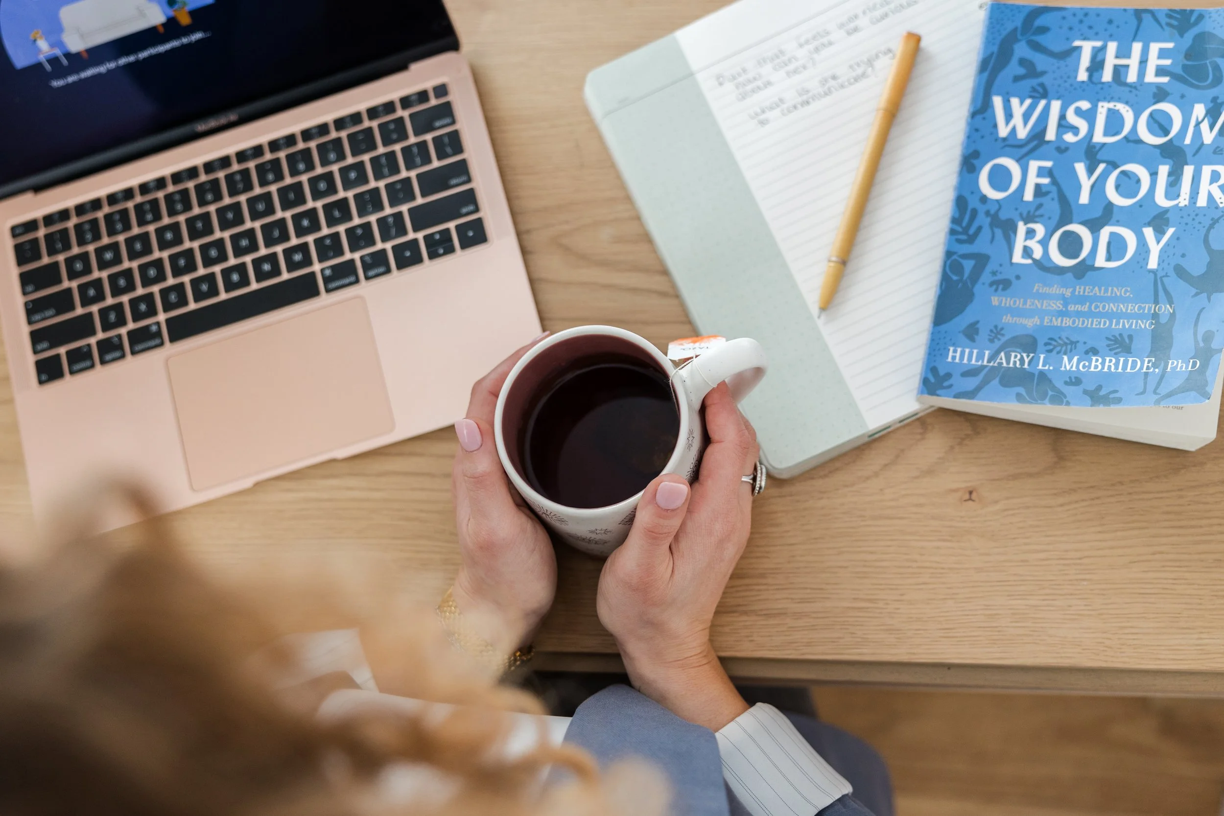 Person holding a mug of coffee or tea in front of an open notebook, with a laptop on the left and a book titled 'The Wisdom of Your Body' on the right on a wooden desk.