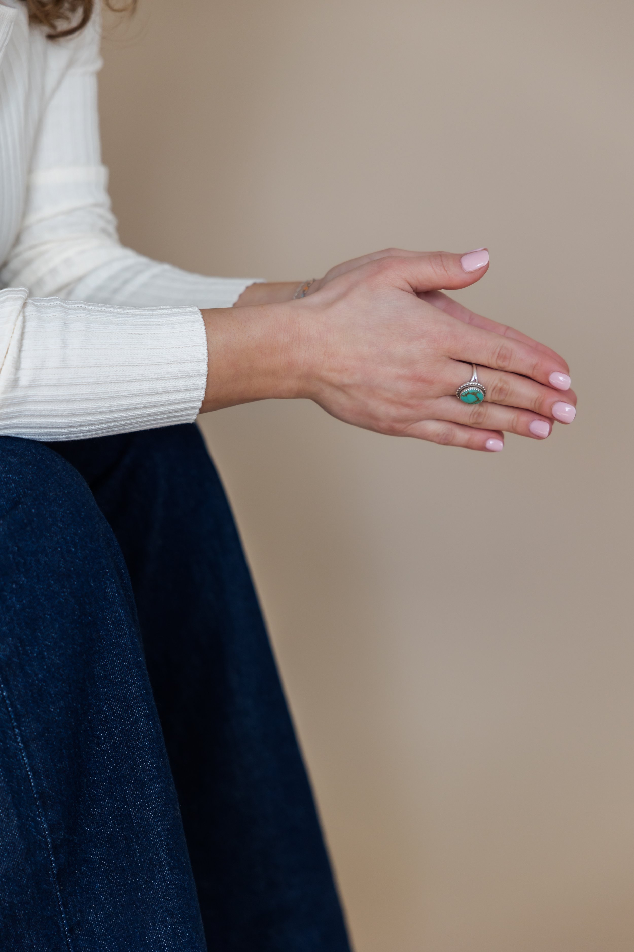 Person with light skin and pink nail polish claps hands together, wearing a white ribbed long sleeve shirt and a turquoise ring, sitting against a plain beige wall.