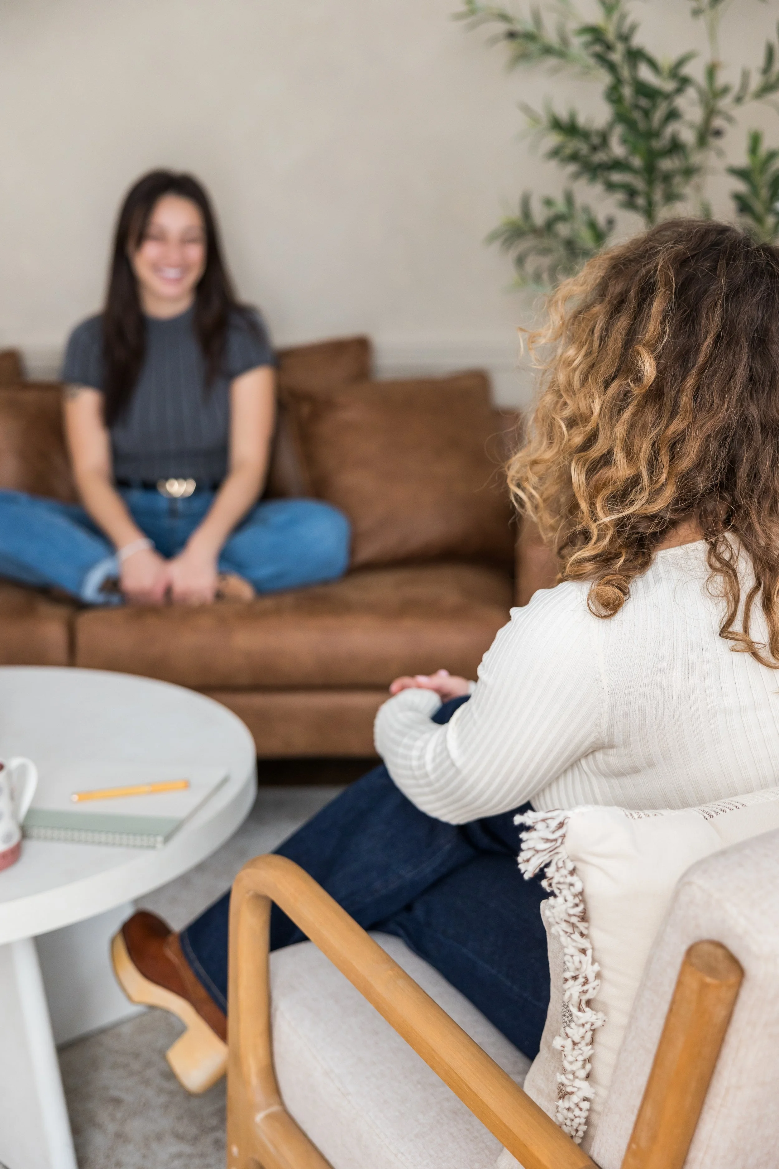 Two women sitting across from each other in a cozy living room, engaged in conversation. One woman with curly hair is seated in a wooden chair, while the other woman with straight dark hair is sitting on a brown couch, smiling.