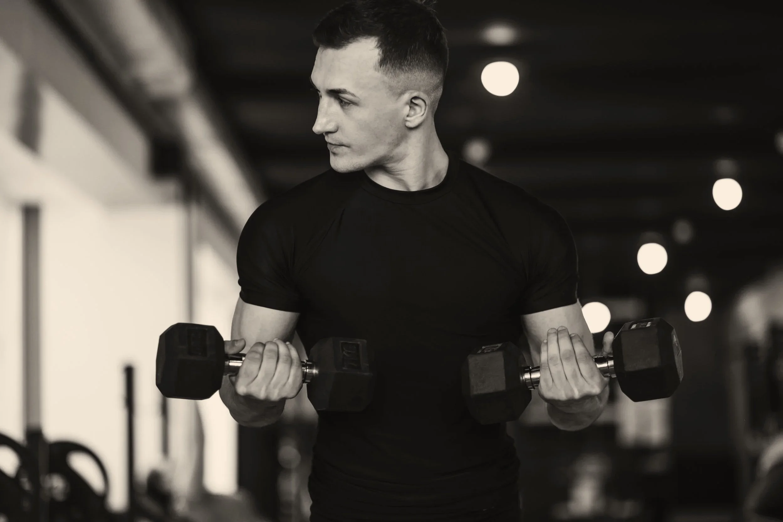 A young man with short hair, wearing a black athletic shirt, lifting dumbbells while exercising in a gym with blurred background lights.