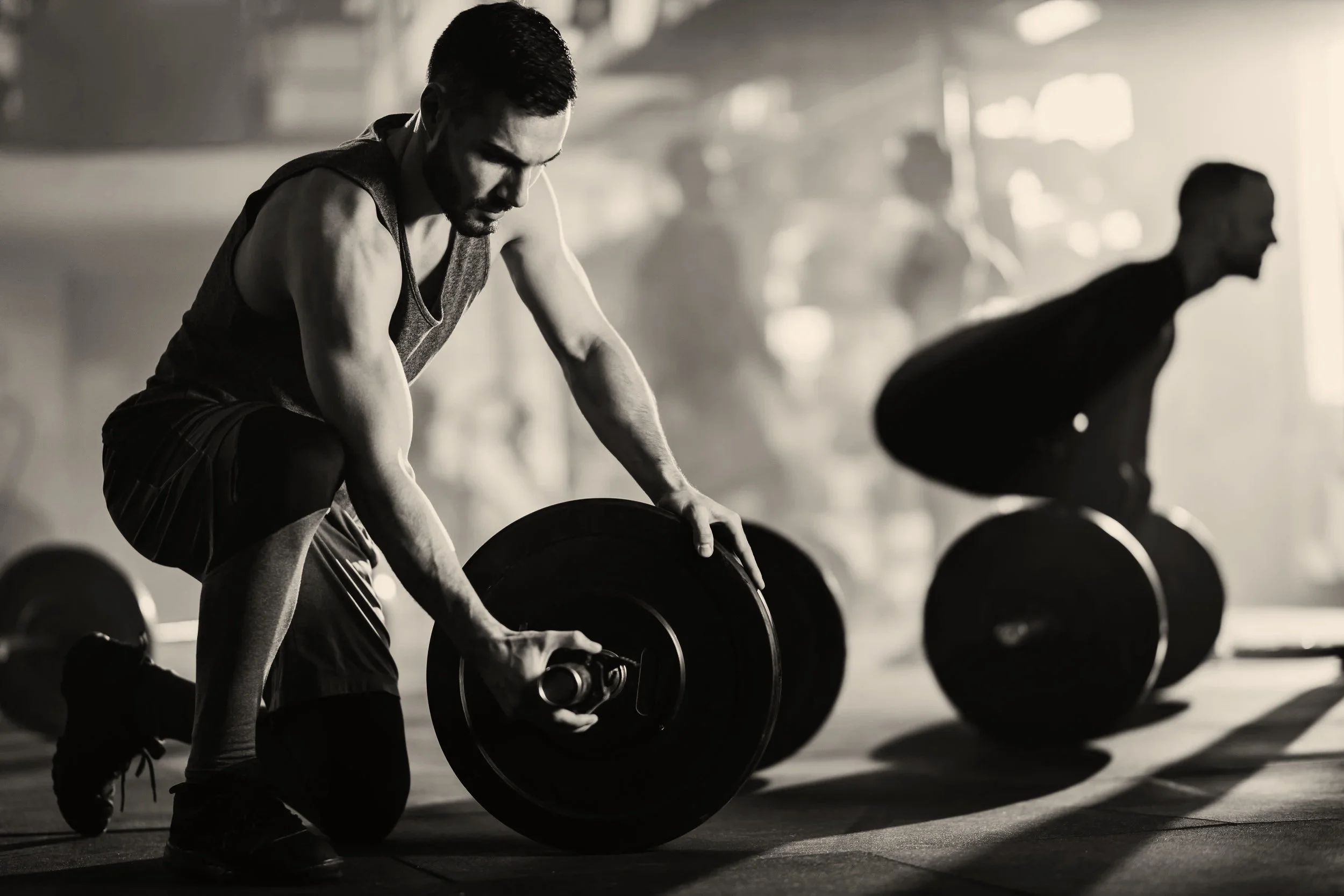 Man in a gym lifting weights with a focused expression, others in the background lifting weights as well, in a dimly lit gym.