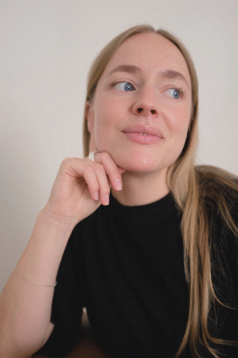 Portrait of a woman with blonde hair and blue eyes, wearing a black top, resting her chin on her hand, looking to the side with a thoughtful expression.