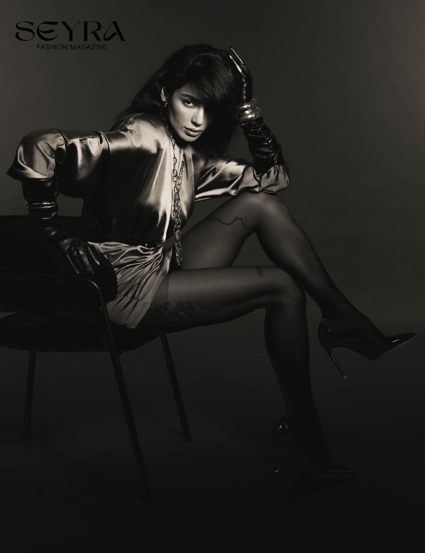 A black and white photo of a woman sitting on a chair, wearing a satin dress, long gloves, high heels, and jewelry. She has dark hair and a confident expression, with her hand resting on her head. The background is plain and dark. The magazine title 'SEYRA FASHION MAGAZINE' is in the upper left corner.