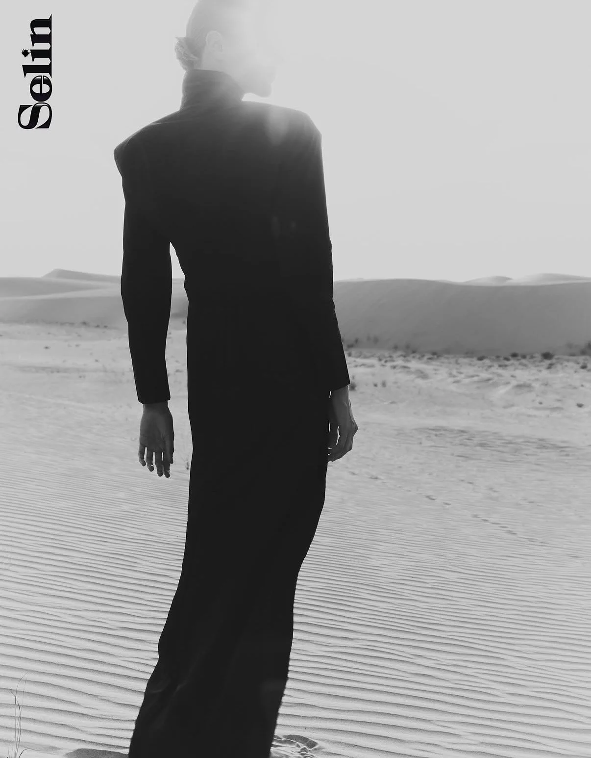 A person in a long black dress walking on sand dunes in a desert landscape during daytime, with sun glare creating a bright halo around their head.