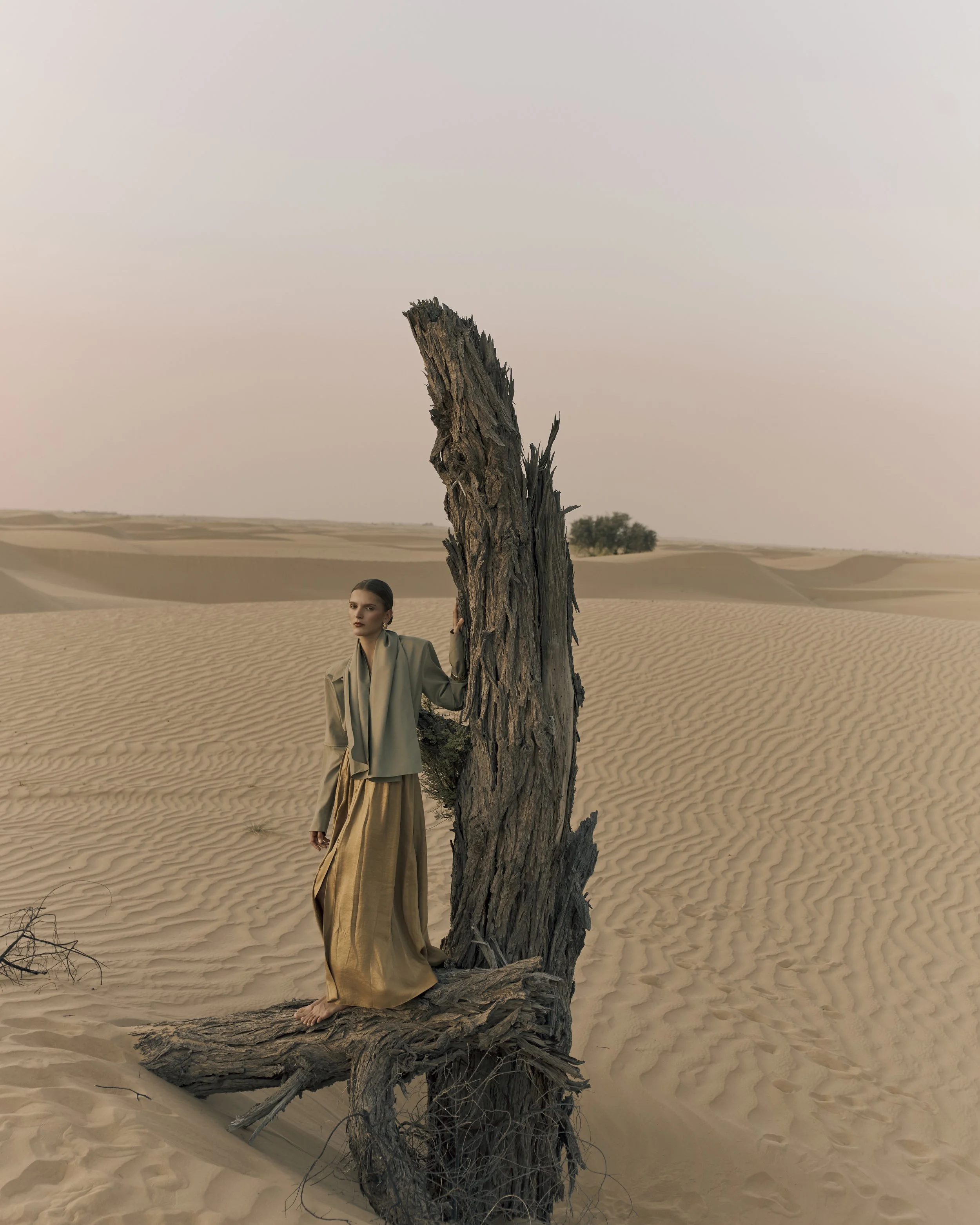 Woman standing in a desert landscape with sand dunes, holding onto a large, weathered piece of driftwood, wearing a beige jacket and long flowing skirt.