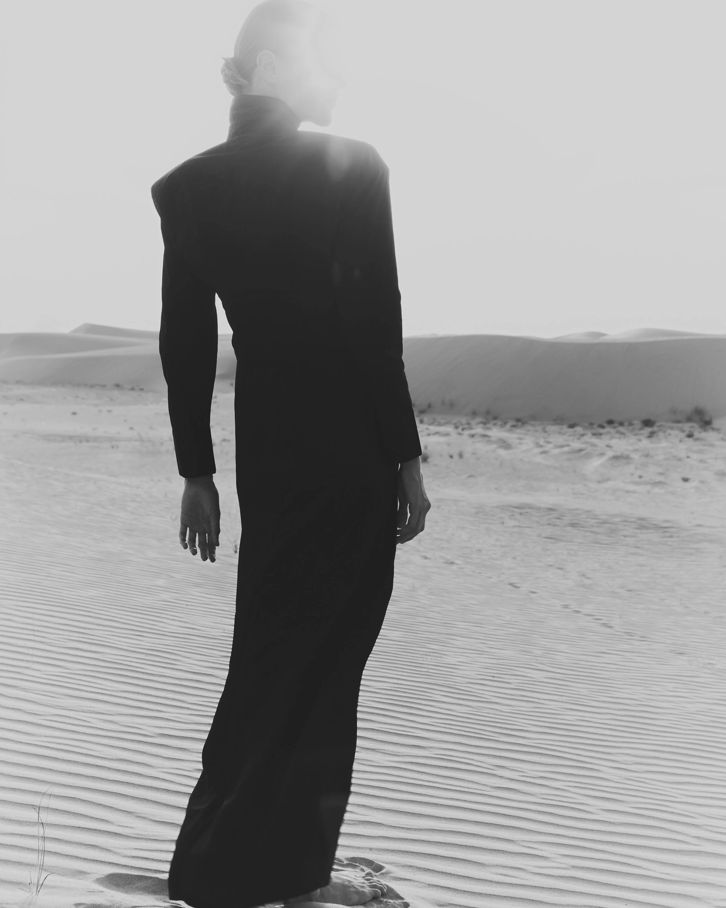 A person with long hair in a bun, wearing a dark jacket and long skirt, kneeling on sand dunes at the beach, facing sideways with sun shining brightly behind; black and white photo.