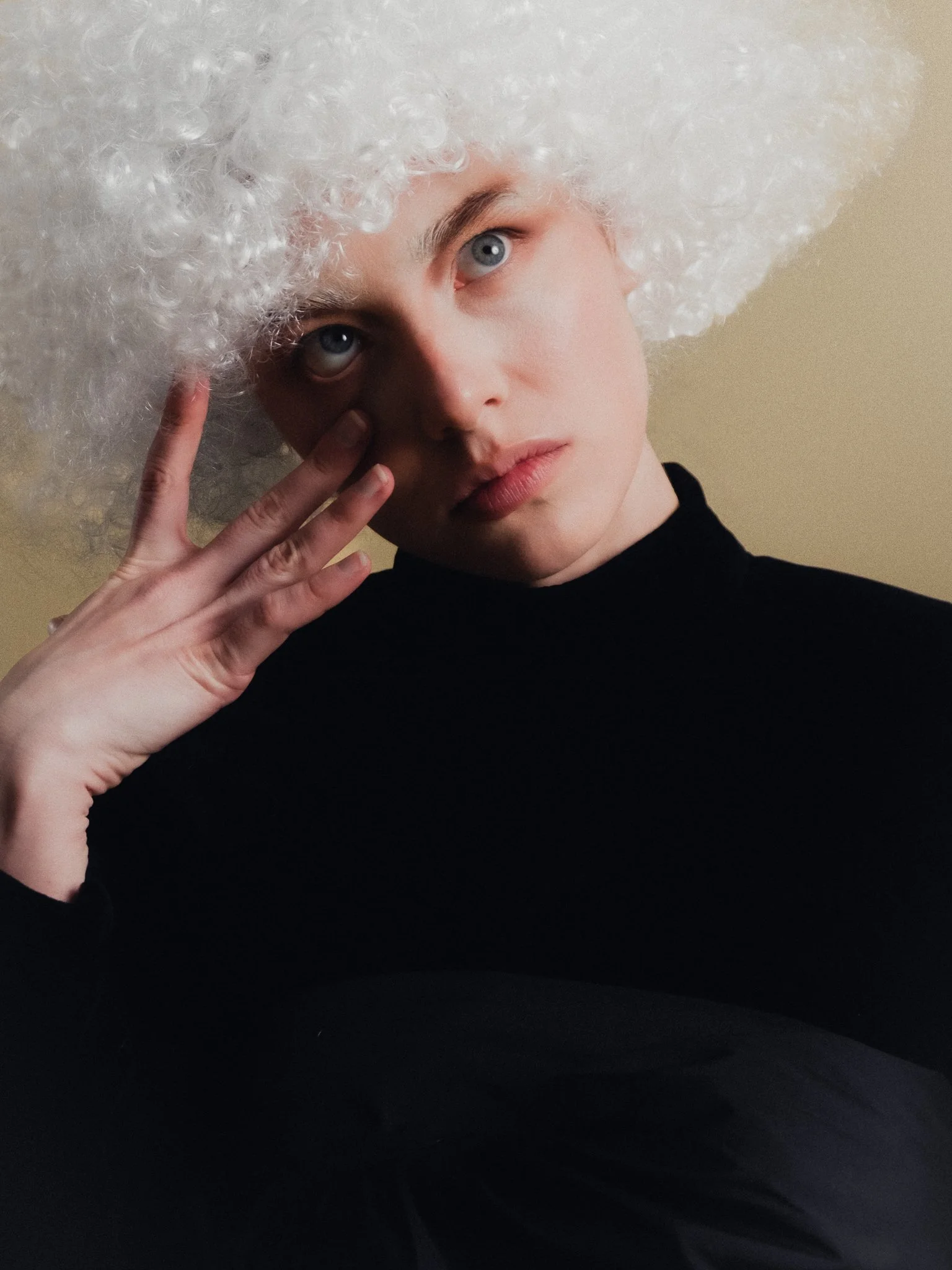 Person with curly white wig and pale skin, dressed in black, touching their face with fingers, looking contemplative.