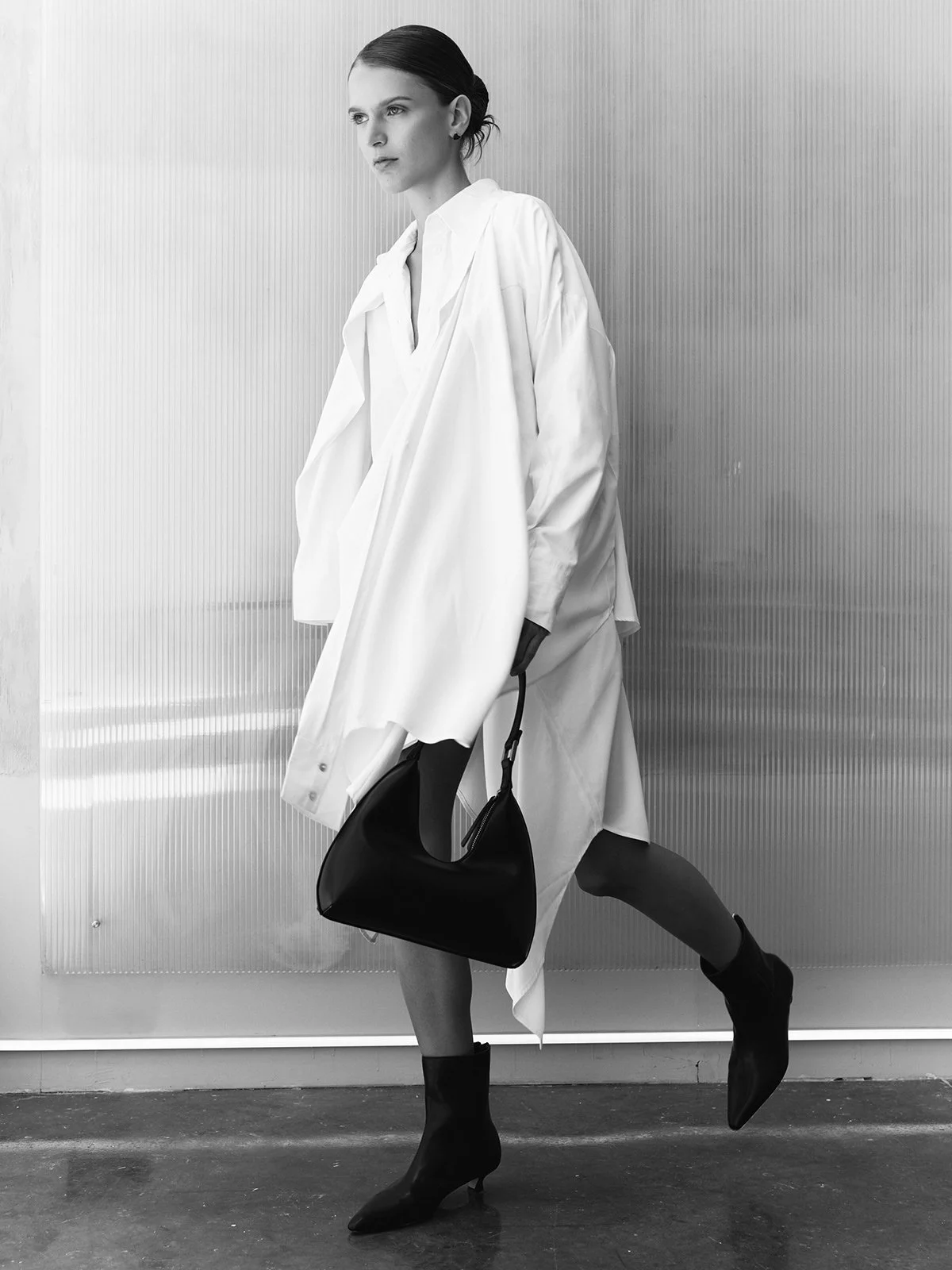 Black and white photo of a woman in a loose white shirt holding a black handbag, standing against a metallic textured wall, wearing black high-heeled boots and posing with one leg crossed in front of the other.