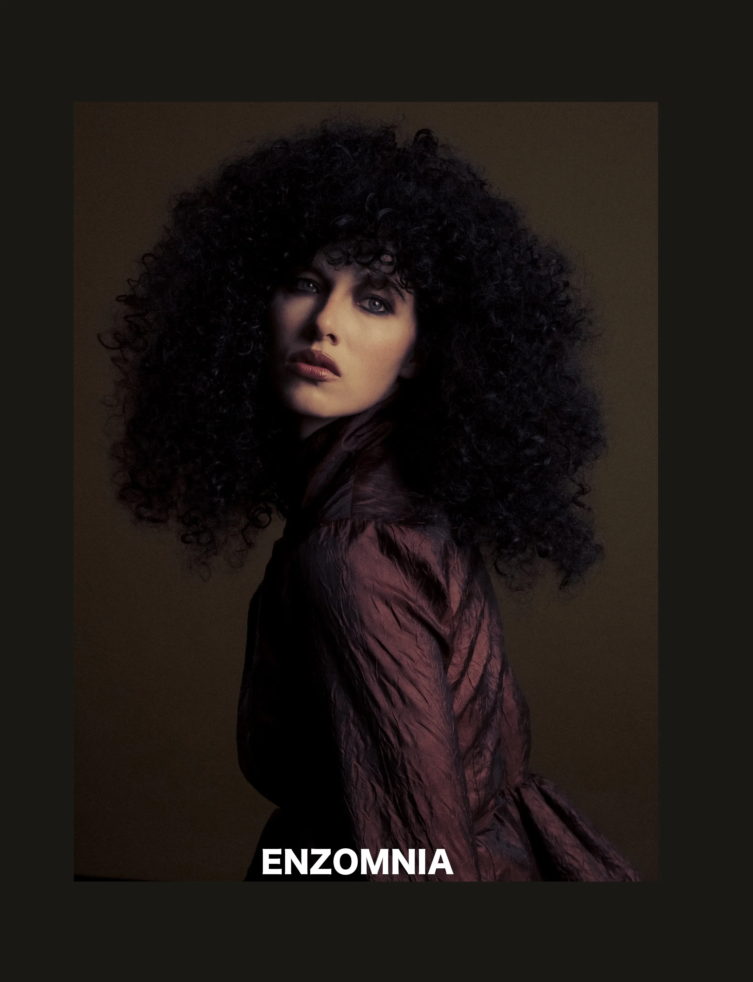 A woman with dark curly hair looking directly at the camera, wearing a dark, textured top, against a brownish background with the text "ENZOMNIA" at the bottom.