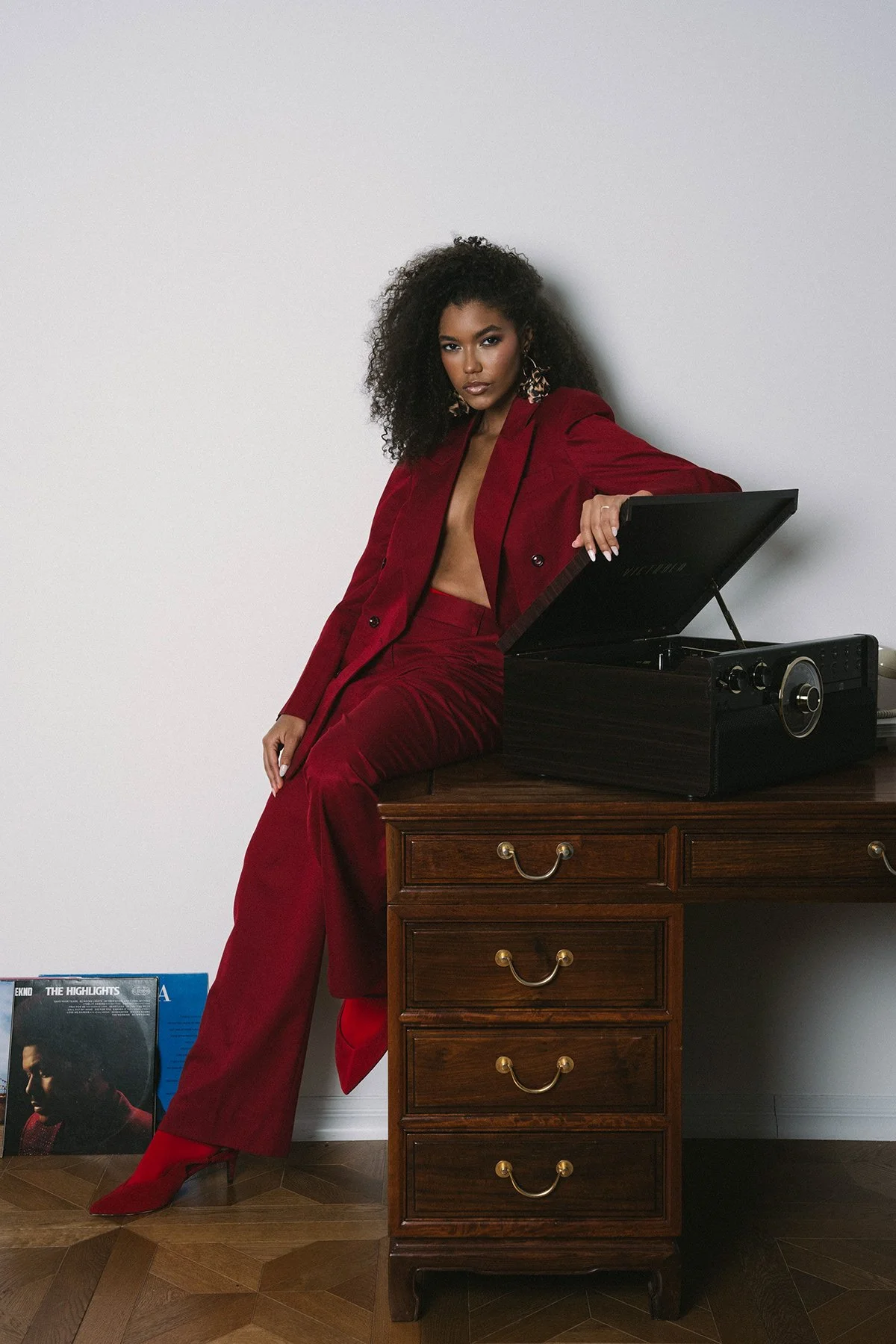 A woman with curly black hair in a red suit and high heels leaning on a wooden dresser with a vintage record player on top. Vinyl records are visible on the floor.