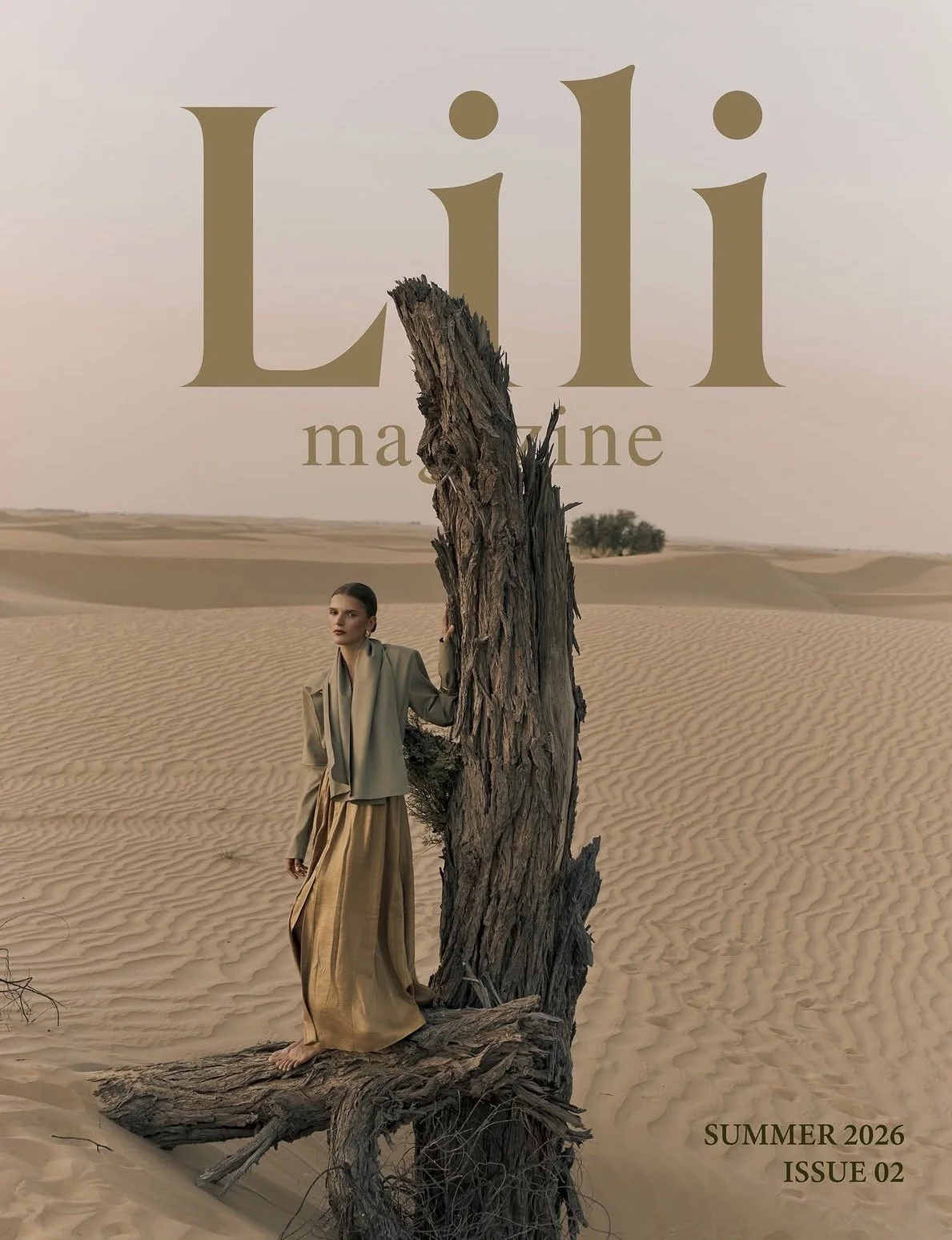 A woman standing in a desert landscape, holding onto a large, weathered tree trunk. In the background, rolling sand dunes and a sparse tree line are visible. Overlaid text reads 'Lili magazine,' 'SUMMER 2026 ISSUE 02'.