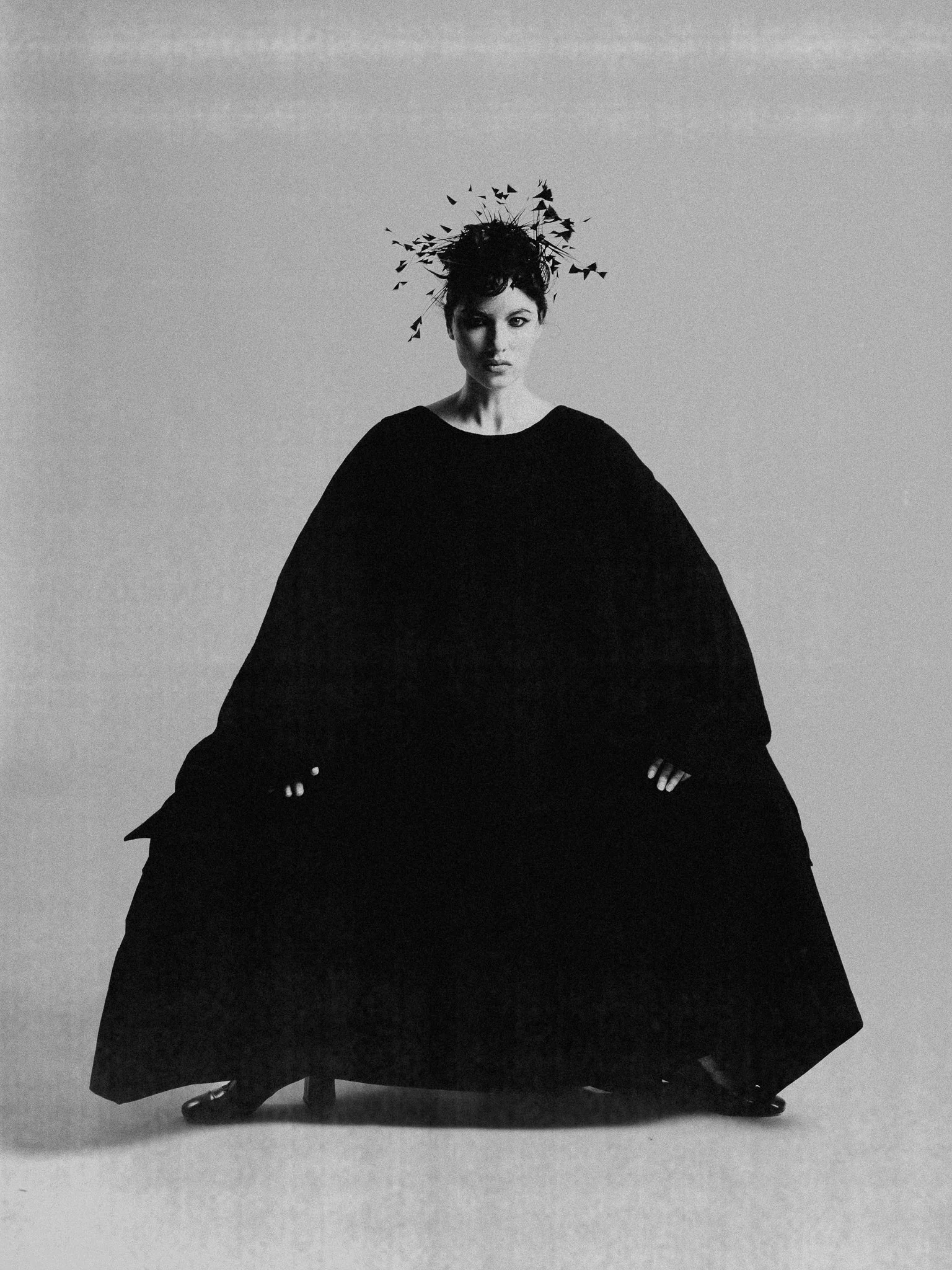 A black and white photo of a woman with short hair, wearing a long flowing black dress with a voluminous skirt, standing against a plain backdrop. The woman's head is adorned with a headdress made of branches and leaves.
